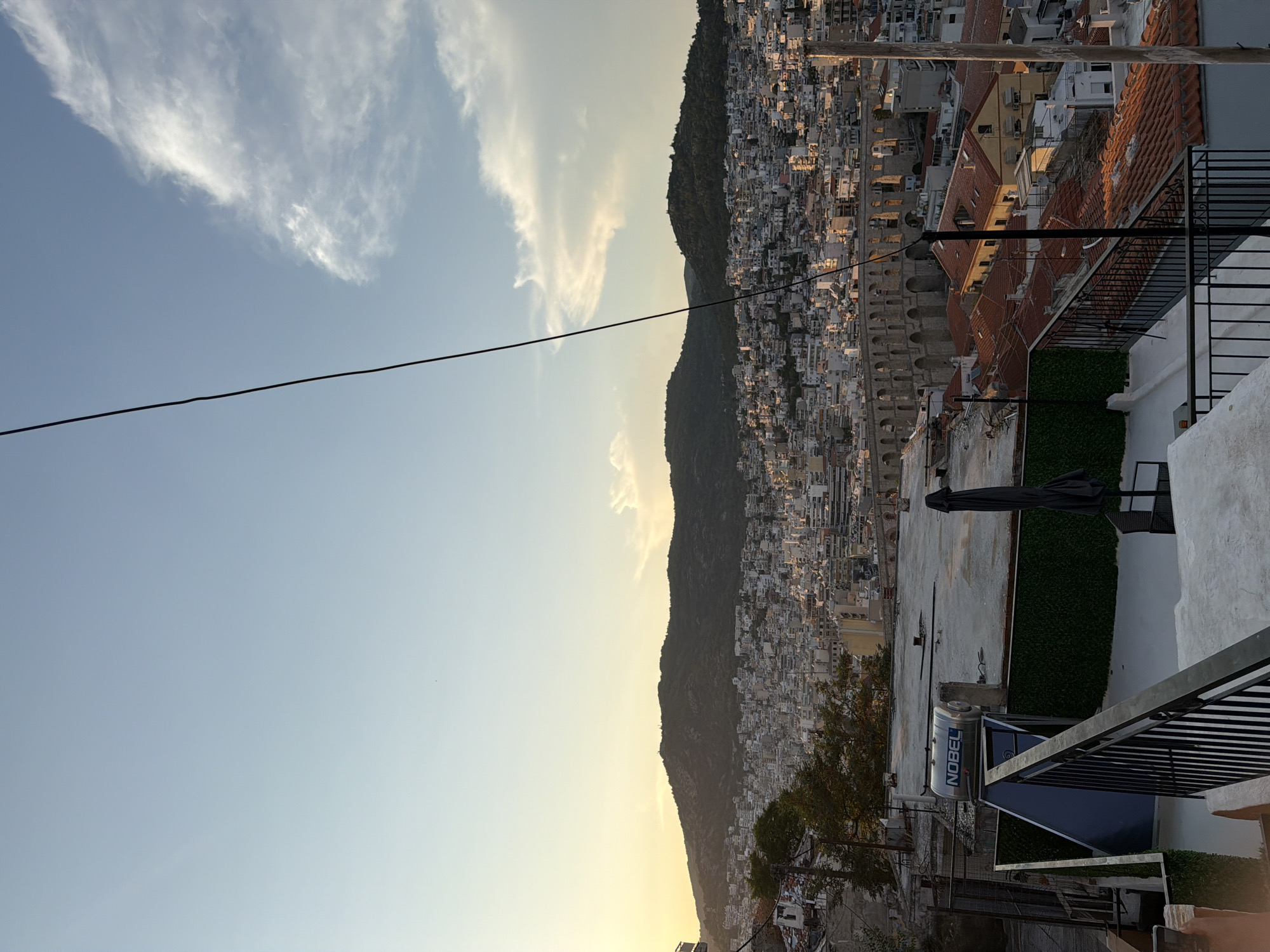 The image captures an elevated view of the city of Kavala, Greece, during what appears to be late afternoon or early evening, given the warm, soft light. In the foreground, a rooftop terrace is visible with a black metal railing. A solar water heater with the brand name "NOBEL" is mounted on the rooftop. A closed black umbrella stands near a table and chairs, suggesting an outdoor seating area.
Beyond the terrace, the cityscape unfolds, characterized by dense clusters of buildings with predominantly white and light-colored facades and red-tiled roofs. The buildings are built on a hillside, ascending towards a prominent, tree-covered mountain range that forms the backdrop.
An impressive ancient aqueduct, a significant landmark of Kavala, stretches across the middle ground, its stone arches clearly discernible. The aqueduct is a key historical feature of the city. The sky above is a soft blue with wispy white clouds, illuminated by the setting sun, which casts a warm glow on the scene. A thin black wire, likely a power line or cable, diagonally crosses the upper portion of the image, extending from the top edge. The overall impression is one of a historic city nestled against a natural landscape, bathed in gentle sunlight.