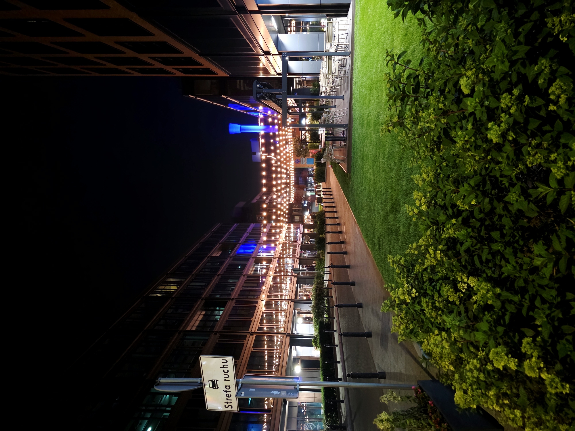 The image depicts an outdoor scene at night in Warsaw, Poland, likely in a modern urban development with commercial buildings and pedestrian areas. On the left, a white traffic sign with a car icon and the text "Strefa ruchu" (Traffic zone) is mounted on a metal pole. Adjacent to the sidewalk, a line of bollards separates the pedestrian area from a grassy lawn.
The buildings on both sides of the street are contemporary structures with large glass facades reflecting the string lights that illuminate the area. These lights are draped across the street, creating a festive atmosphere. In the foreground, to the right of the grassy lawn, there are several outdoor tables and chairs, suggesting a restaurant or cafe seating area. The lawn is neatly manicured and illuminated, making it stand out against the dark sky. In the far background, a tall, blue-lit cylindrical structure is visible. The overall scene is one of an urban commercial district after dark, with decorative lighting enhancing the ambiance of the street and outdoor dining spaces. There are no people visible in the image.