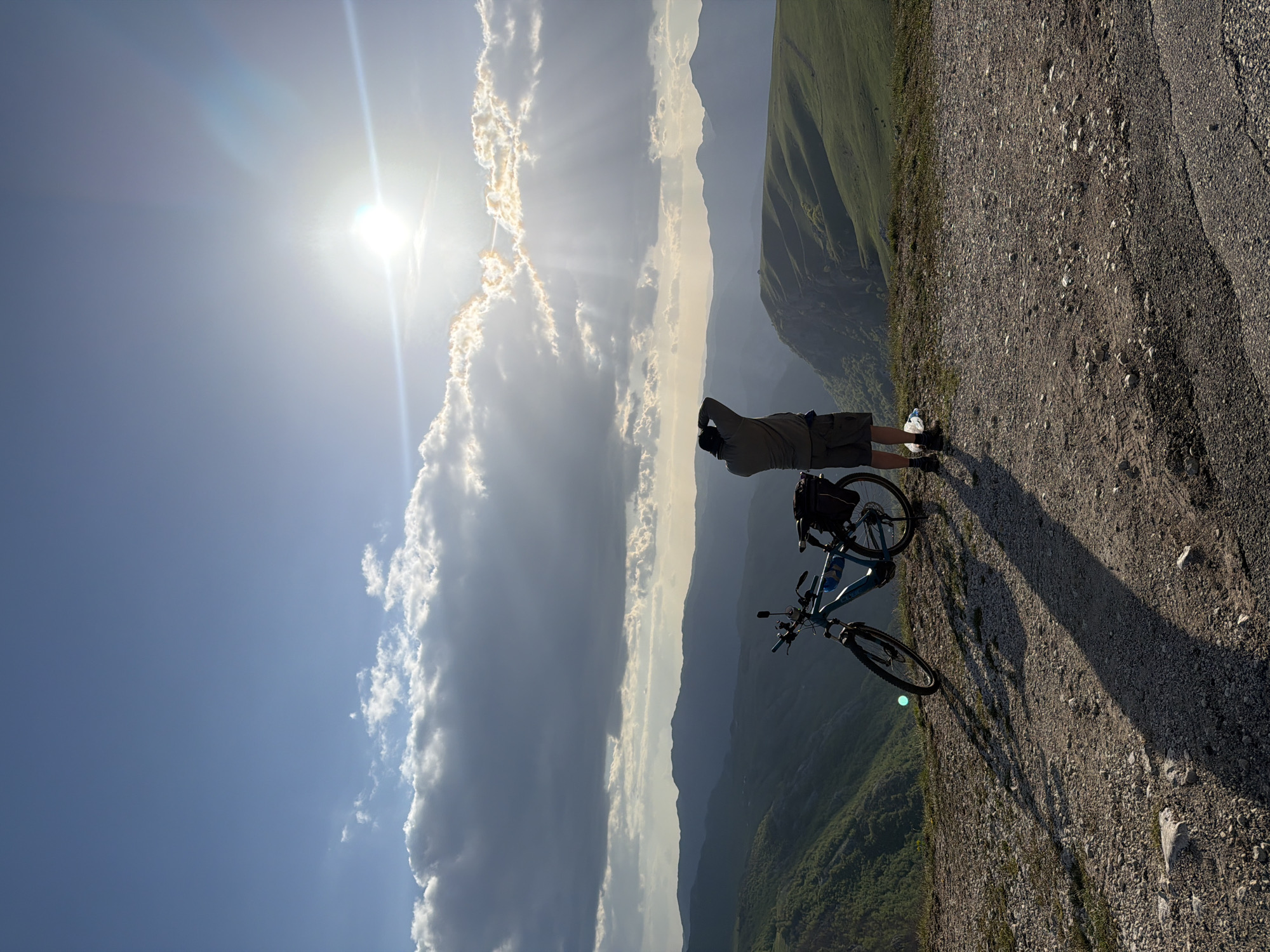 A man stands with his bicycle on a gravel path, observing a mountain range under a bright, sunny sky with large cumulus clouds. The man is wearing shorts and a light-colored shirt and is shielding his eyes with his hands. His bicycle is light blue. The location appears to be at a high elevation, near Pišče, Montenegro, evidenced by the mountainous terrain. The gravel path is uneven, suggesting a remote, unpaved road.