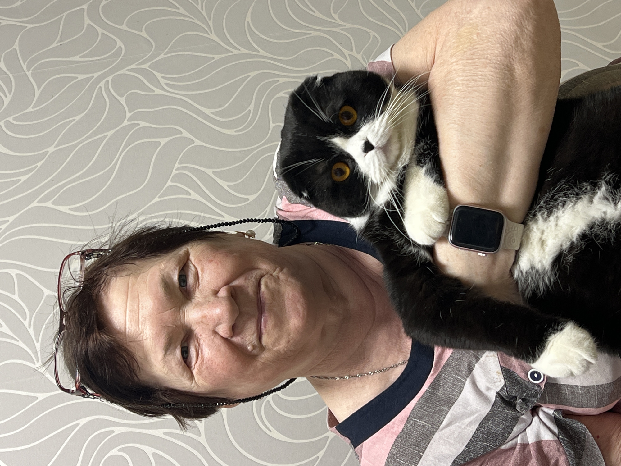 An adult woman is holding a black and white cat. The woman has short brown hair and is wearing glasses. She is wearing a striped shirt. The cat has yellow eyes and white whiskers. The woman is wearing a smart watch on her wrist. The background is a gray wall with a white wave pattern. There is no indication that this image was taken in Perm, Russia.