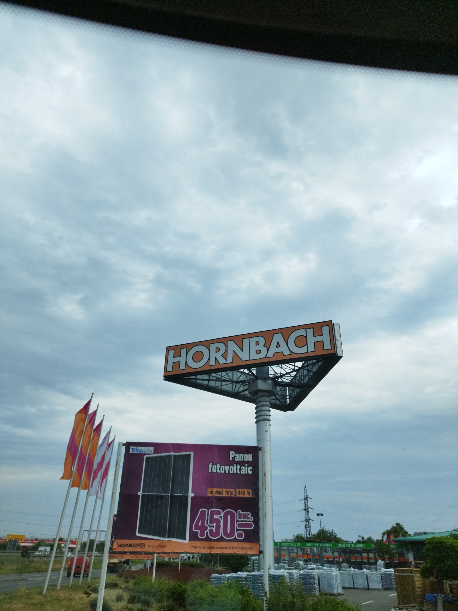 A large, triangular HORNBACH sign is mounted on a metal pole against a cloudy sky. Below the sign, a billboard advertises "Panou fotovoltaic" (photovoltaic panel) for 450 lei, with a "TrinaSolar" logo and an image of two solar panels. Several colorful flags, orange, pink, and white, are displayed on poles to the left of the billboard. In the background, a road with a red car is visible, along with other buildings and a power line tower. The scene appears to be in an outdoor commercial area, possibly a parking lot or storefront, with stacks of goods and greenery. The location is identified as Domnești, Romania.