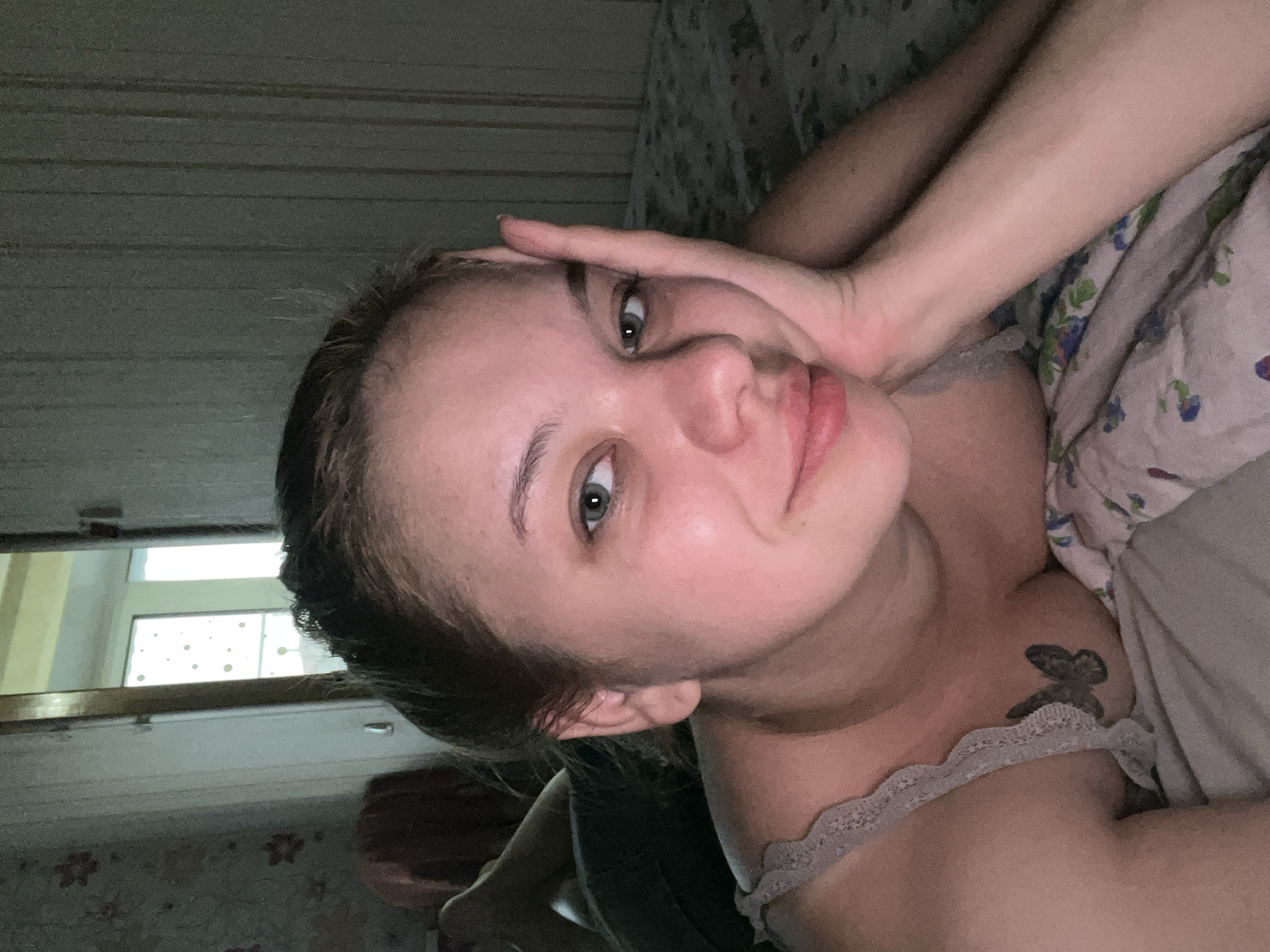 This media file features a young woman lying in bed, taking a selfie. She is positioned on her side, with her head tilted and her left hand resting on her forehead. Her eyes are open and looking towards the camera, and she has a neutral expression. She is wearing a light-colored top with lace trim. A tattoo of a butterfly is visible on her left shoulder.
The setting appears to be a bedroom. Behind her, the wall has a horizontal striped pattern. To her left, a window is visible, reflecting light. The overall lighting suggests it might be late evening or early morning. The scene is intimate and personal, with the subject in a relaxed posture. No text is visible in the image. The location context is Gorodets, Russia.