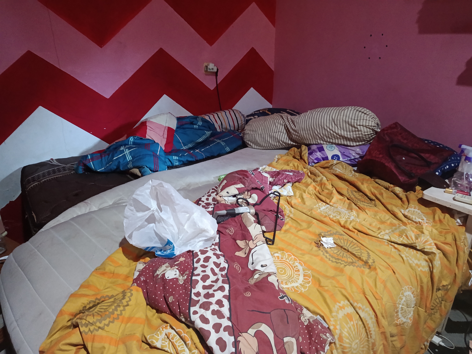 The image depicts a messy bedroom. A bed is covered with various blankets and pillows, some with patterned designs. A white plastic bag is placed on the bed. A wall behind the bed is painted with a red and pink zigzag pattern. A light switch and electrical outlet are visible on the wall. A small table with some items on it is present in the corner of the room. The scene appears to be in a residential setting in Keboananom, Indonesia.