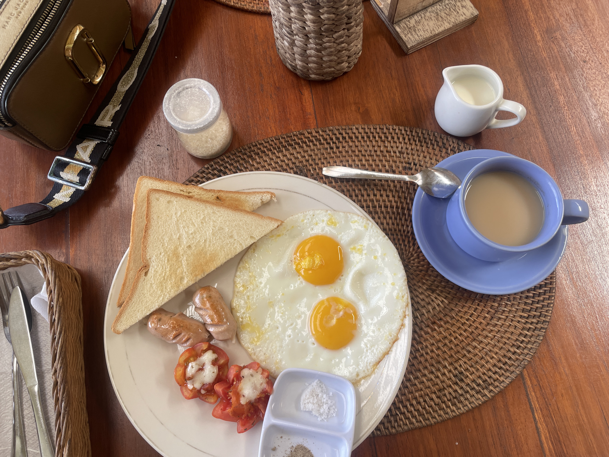 A top-down shot reveals a breakfast setting likely within the Ubud District of Indonesia. A white plate displays two fried eggs with runny yolks, alongside two slices of toasted bread, two sausages, and tomato slices topped with a white substance, potentially cheese. A small condiment dish containing salt and pepper sits on the plate. A blue teacup filled with a light-colored liquid, possibly tea or coffee with milk, rests on a matching saucer. A small white creamer pitcher is nearby. To the left, a woven basket holds silverware wrapped in a white napkin. A small jar with a clear lid containing granulated sugar is also present. A woman's purse with a decorative strap sits to the left of the plate. The entire arrangement is placed on a circular woven placemat set on a brown wooden table. Other decorative items are visible in the background, adding to the ambiance.