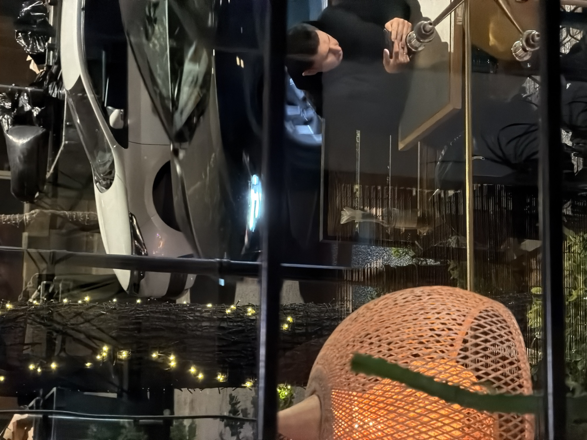 The image depicts a complex scene captured through a glass partition, resulting in a blend of interior and exterior elements, likely during the evening or night in Yerevan, Armenia.
In the upper central portion of the image, the reflection of an adult male is visible. He has short dark hair and is wearing a dark, long-sleeved top. He is looking down intently at a smartphone held in both hands. Parts of what appear to be interior furnishings, including a table and metallic decorative elements, are also reflected or visible in the background behind him.
Reflected on the left side of the glass, a white car, possibly a modern electric vehicle, is seen upside down. Nearby, black plastic bags or covers are draped over an unseen object or structure.
The foreground of the image features a large, intricately woven wicker-style lamp or decorative light fixture in the bottom right corner, emitting a warm, orange glow. Across the lower half of the image, numerous small, yellow fairy lights are strung along a dark, textured horizontal surface, possibly a railing or decorative element, adding to the ambient lighting. Vertical and horizontal structural elements, likely window frames or architectural beams, crisscross the view. In the deeper background, through the glass, hints of green foliage and a decorative wall with subtle vertical patterns can be seen. There is no visible text in the image.