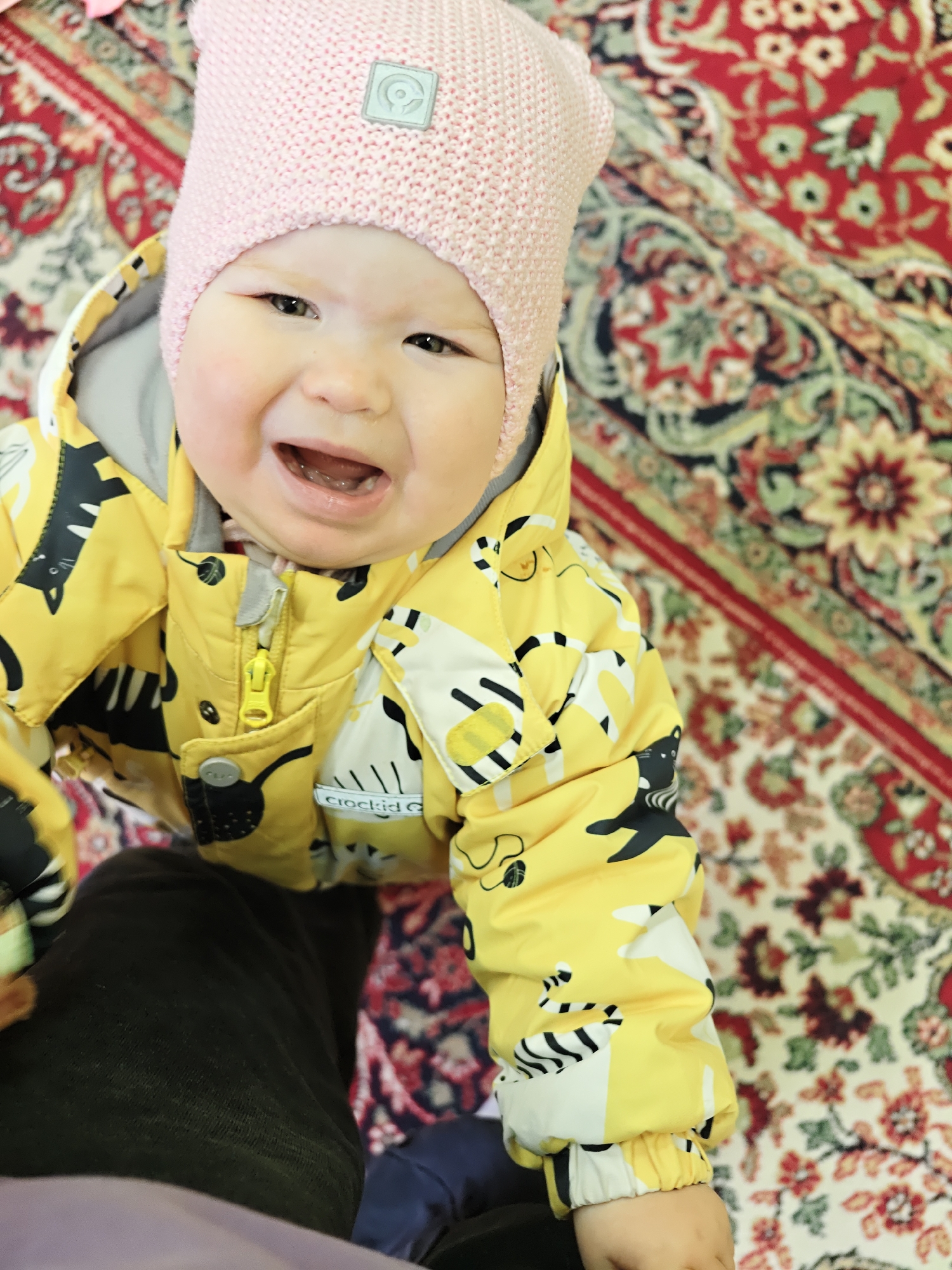 A baby wearing a pink knit hat and a yellow jacket with a pattern of black and white cats stands on an ornate rug. The baby appears to be crying or smiling widely, with mouth open. The rug has a floral pattern in red, beige, and green. A person's dark pants leg is visible in the lower left of the frame. This photograph may have been taken in Chaikovsky, Russia.