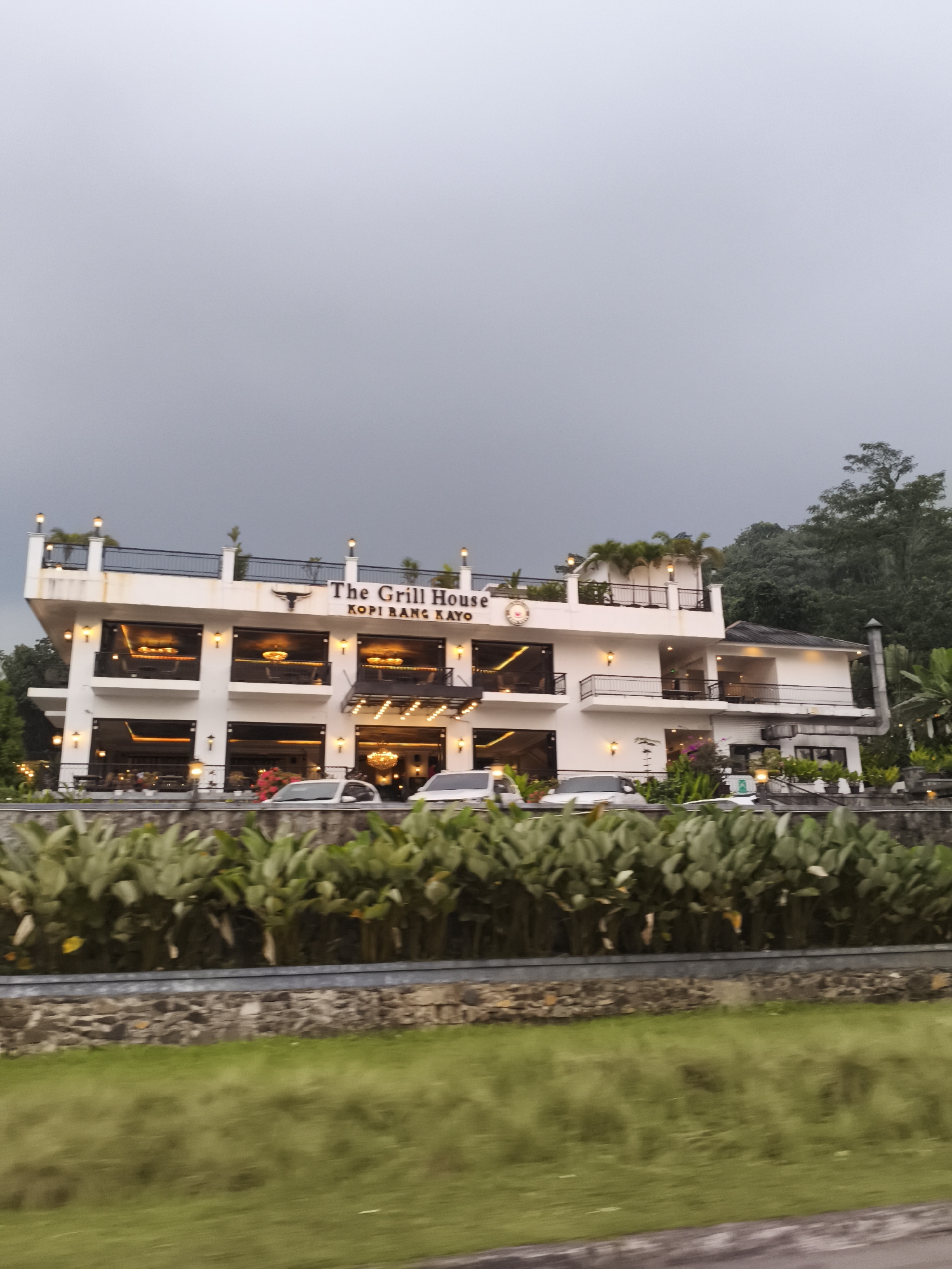This media file captures a roadside view of a prominent two-story building identified as "The Grill House KOPI BANG KAYO," likely a restaurant or cafe, in Babakan Madang, Indonesia. The building is painted white with large, dark-framed windows and balconies, and is brightly illuminated by numerous warm-toned artificial lights along its facade and on its upper terrace. A distinct bull's head logo is visible on the front of the building.
The scene depicts the establishment during what appears to be evening or dusk, given the dependence on artificial lighting and the overcast, grey sky overhead. The interior spaces visible through the windows also show elaborate chandeliers and lighting, suggesting an upscale or inviting ambiance. Several cars are parked in front of the building, hinting at ongoing business.
In the foreground, a blurred strip of green grass and a low barrier with a stone wall separate the viewer from a dense row of green, leafy plants. The blurring effect in the foreground suggests the image was captured from a moving vehicle. Beyond the building, a dense backdrop of dark green trees indicates a natural, possibly hilly or mountainous, landscape, characteristic of areas like Babakan Madang. No people are distinctly visible in the frame. The overall impression is that of a well-lit and active commercial establishment situated within a natural setting.