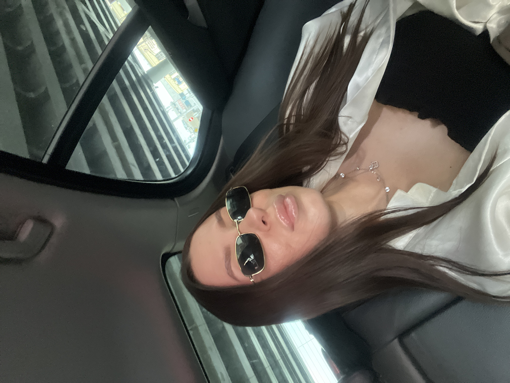 A woman with dark hair is sitting in a car, wearing sunglasses, a black top, and a white shirt. She is wearing a necklace. The car is in a parking garage, likely in Moscow, Russia, given the architectural style visible through the car window.