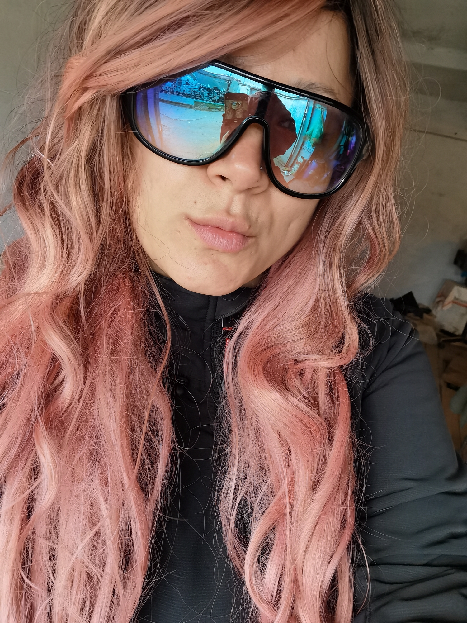 A selfie of a person with long, wavy, pink hair wearing large, reflective sunglasses. The sunglasses reflect a blue and white scene, possibly of a vehicle and a building. The person is wearing a black jacket with a red detail near the neckline. The background appears to be an interior space with miscellaneous objects and boxes. There is insufficient detail to determine the location as Chaikovsky, Russia.