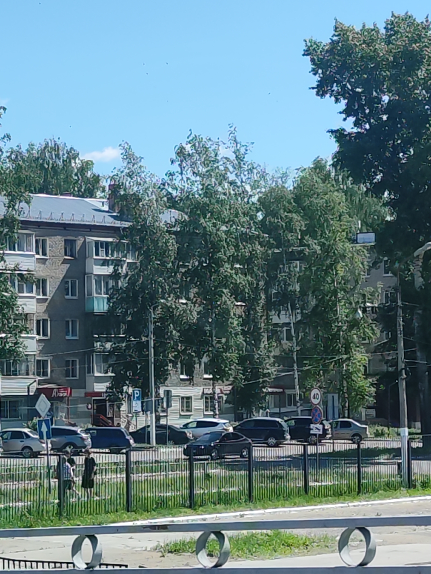 A street view in Chaikovsky, Russia, on a clear, sunny day shows a multi-story apartment building on the left, with several trees in front of it. A paved road with cars is visible in the midground, and a metal fence with grass in front of it is in the foreground. Two people, one wearing a dark dress and the other in lighter clothing, are walking along the sidewalk behind the fence. Traffic signs, including a speed limit sign indicating 40, are present.