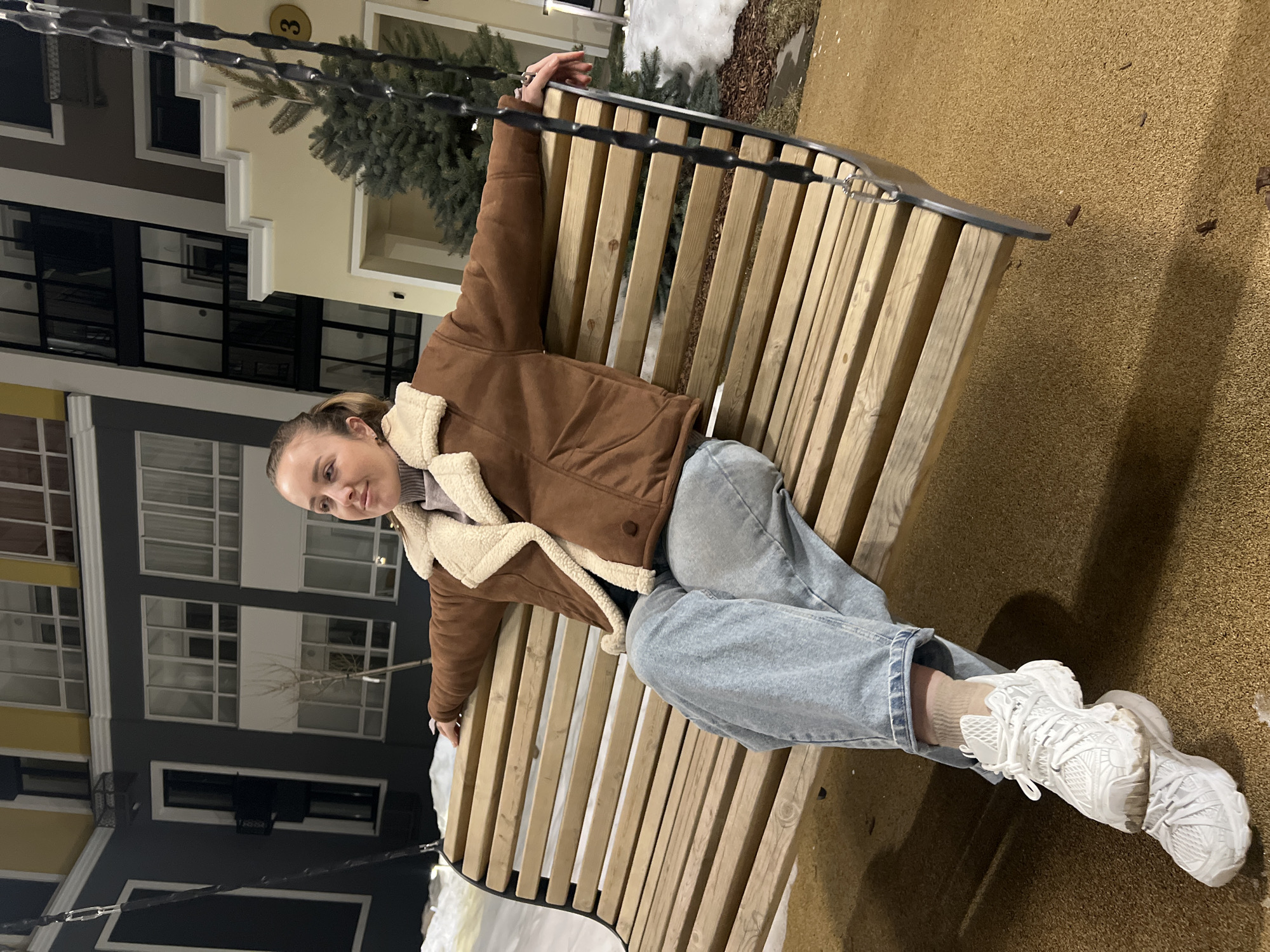 A light-skinned female individual is seated on a wooden slatted swing in an outdoor residential courtyard in Unknown, Unknown. She wears a brown shearling-style jacket with cream fleece lining, a light purple/grey turtleneck sweater, light blue wide-leg jeans, and white chunky athletic sneakers over tan socks. Her hair is pulled back, and her head is turned to her left, looking upwards with a slight smile. Her arms are extended and resting on the outer edges of the swing's backrest. Her left foot is positioned atop her right foot, both resting on the ground.
The swing features dark metal supports and chains with rubberized sections. The ground surface is a granular, tan-colored material, consistent with playground safety surfacing. Patches of snow are visible on the ground to the left and behind the swing.
In the background, multiple modern multi-story residential buildings are present. The building to the left is dark grey with white window frames and a yellow top section. To the right, a lighter yellow/beige building with white trim, dark windows, and a ground-floor entrance is visible. A circular gold plaque with the black numeral "3" is affixed to the upper facade of this building. Evergreen trees and bare deciduous trees are interspersed throughout the scene. The area is illuminated by artificial lighting, suggesting nighttime or late dusk conditions.