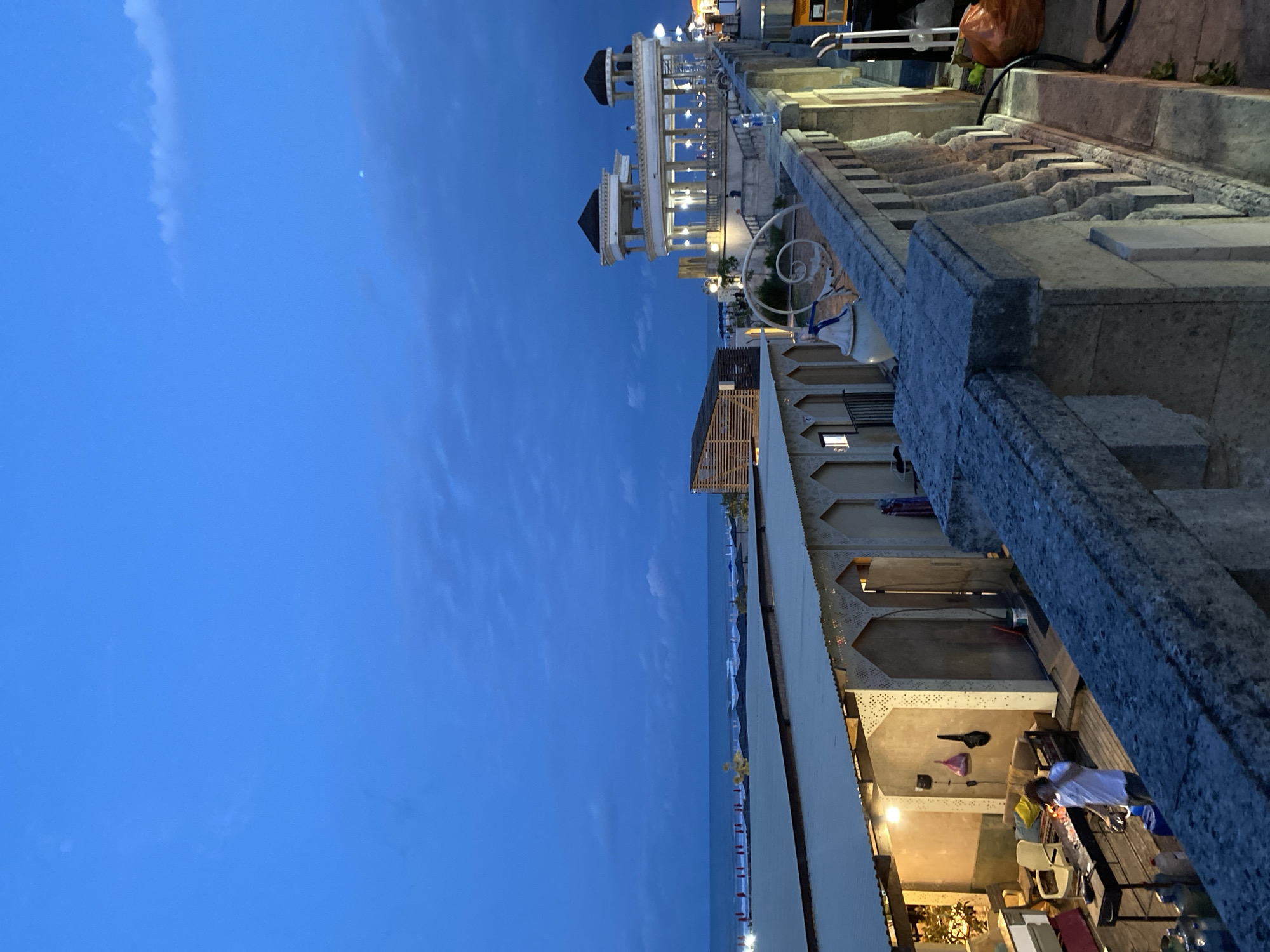 The image depicts a twilight scene at a coastal resort area, likely located in Derbent, Russia. In the foreground, a stone balustrade with ornate carvings lines a walkway. Beyond the balustrade, a low-lying structure with a corrugated roof and arched openings is visible. Inside this structure, a person in a white t-shirt and dark pants is seen standing near a grill, possibly engaged in cooking or preparation. Several gas cylinders are present near the grill. Further in the background, the Caspian Sea stretches out, with gentle waves visible. On the right side of the frame, a multi-tiered white building with columns and a dome-like roof stands illuminated by lights, suggesting it may be a restaurant or a pavilion. The sky above is a deep blue, with some scattered clouds, and a faint hint of the moon is discernible. The overall impression is one of a leisurely evening by the sea, with a mix of architectural styles and recreational activities.