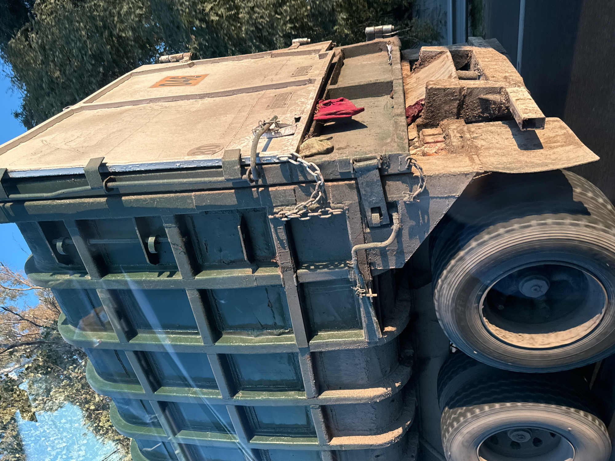 The image shows a close-up view of the top of a truck or trailer, possibly viewed from another vehicle through a window (suggested by reflections). The truck's upper section is largely green, with a grid-like structure. The top of the truck is flat, covered in a light-colored material with what appears to be a red cloth or object on top. Chains are visible securing the top. The trailer has four tires and there is a road in the background. The location is unknown. The photograph does not contain any evidence to suggest that it was taken in the city of Buenos Aires, Argentina.