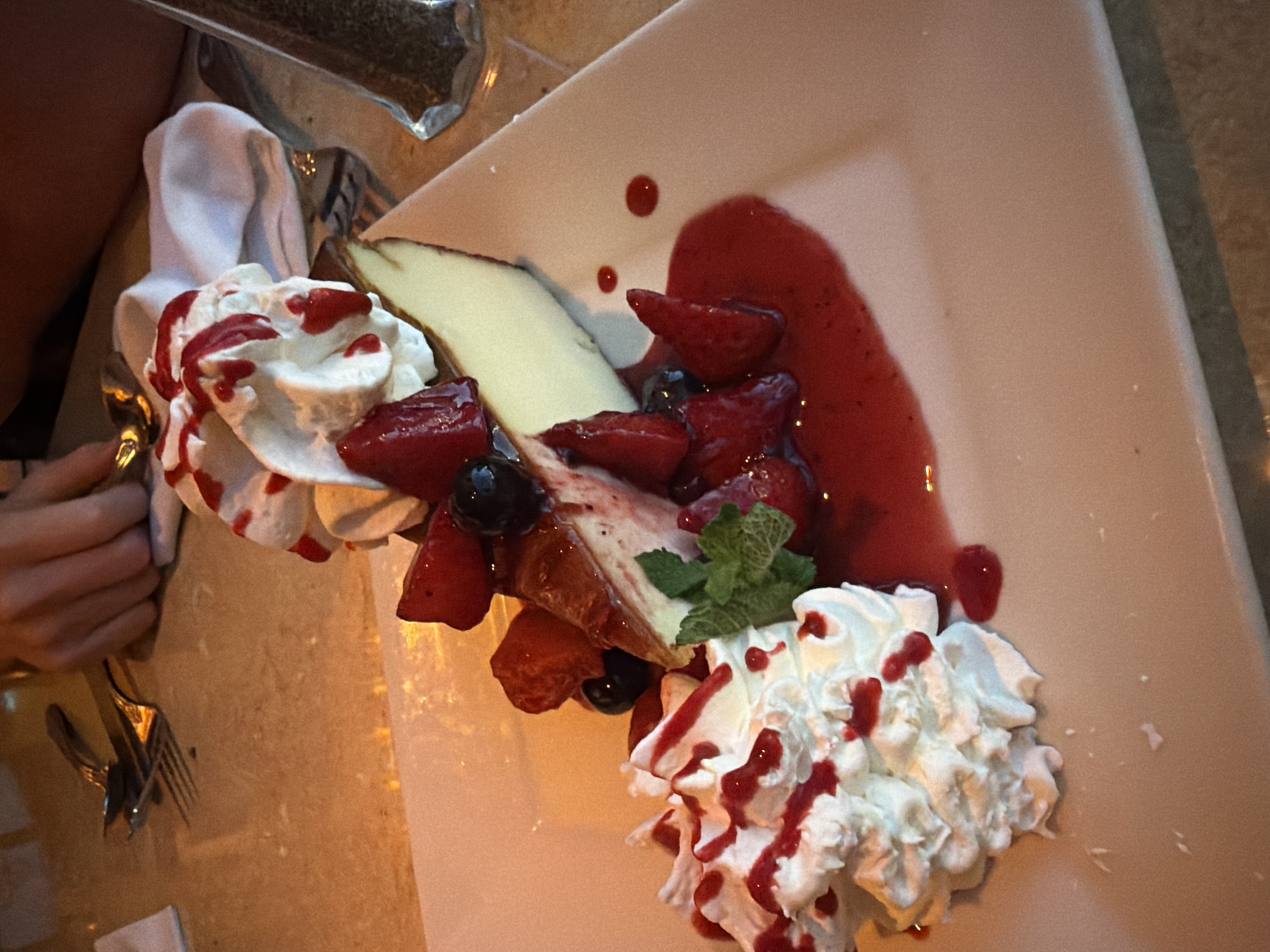 A slice of cheesecake topped with whipped cream, strawberries, and blueberries sits on a white plate. The plate is on a table with a marble-like surface. In the background, a person's hands are visible, holding silverware. The lighting suggests an indoor setting, possibly a restaurant. There is no indication of the city or country.