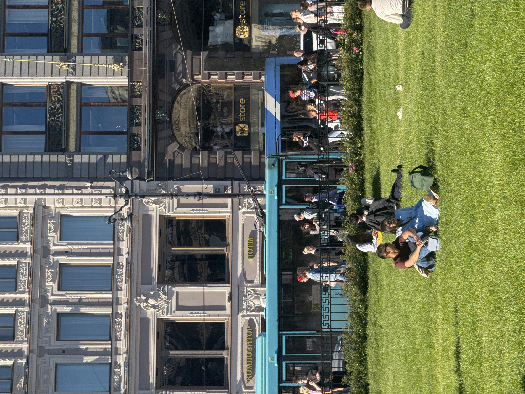 The image depicts a sunny day in Saint Petersburg, Russia, likely on Nevsky Prospekt, given the architectural style and the presence of a tram. In the foreground, two individuals are seated on a lush green lawn, facing away from the camera, with their legs crossed. A green backpack rests on the grass beside them, and a light blue bag is placed between them. Further back, a turquoise tram with white stripes moves past a decorative black fence. A diverse group of people are visible on the sidewalk behind the fence, some standing and conversing, others walking. The background is dominated by the ornate facades of classical European-style buildings. On the left, a building with light-colored stone and classical detailing features multiple windows with light curtains. To its right, a darker building with a grand arched entrance and shop windows with gold lettering that reads "VK store" is prominent. The sky is clear blue.
