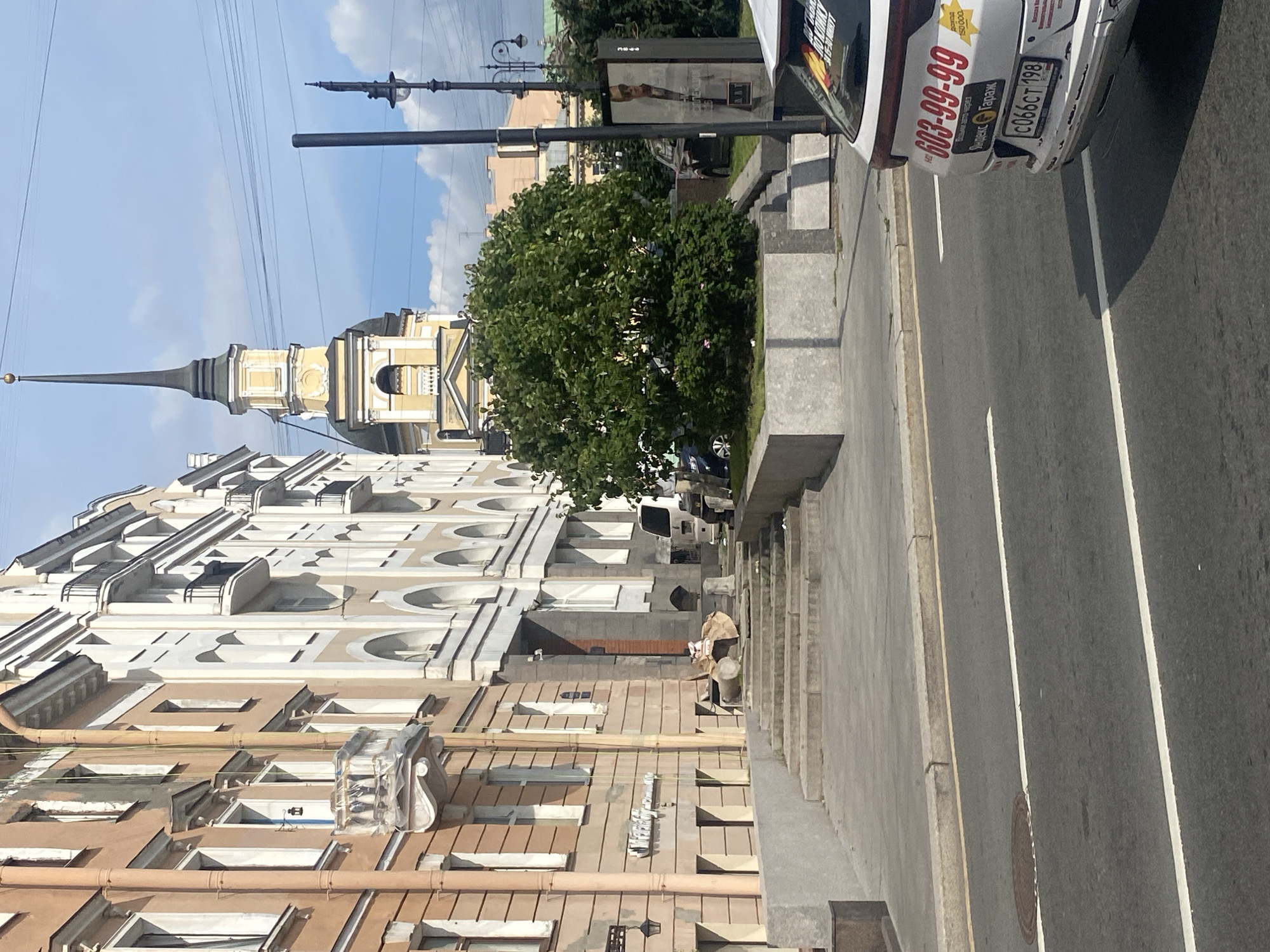 The image captures a street scene in Saint Petersburg, Russia. In the foreground, a white car with a license plate reading "C066CT 198" and prominent red lettering "603-99-99" is parked on the right side of the road. The car has stickers on its rear, one yellow with red text and another red and yellow sticker. The street itself is asphalt with white lane markings and a manhole cover visible.
To the left of the street, a series of concrete steps lead up to a building entrance. The building on the left is an apartment building with light brown and cream-colored facades, featuring numerous windows and balconies. Some balconies are covered with plastic sheeting, suggesting ongoing renovation or protection. To the right of the steps, a large, dense green tree is planted in a raised concrete planter.
Behind the tree, a tall, slender church tower with a yellow facade, dark dome, and a pointed spire topped with a golden ball dominates the background. The sky is blue with scattered white clouds. Further in the background, other buildings and utility poles are visible. A streetlamp stands near the church tower. A billboard featuring a portrait of a man in sunglasses is positioned behind the tree, to the right of the church. The overall impression is a typical urban street in a historical district.