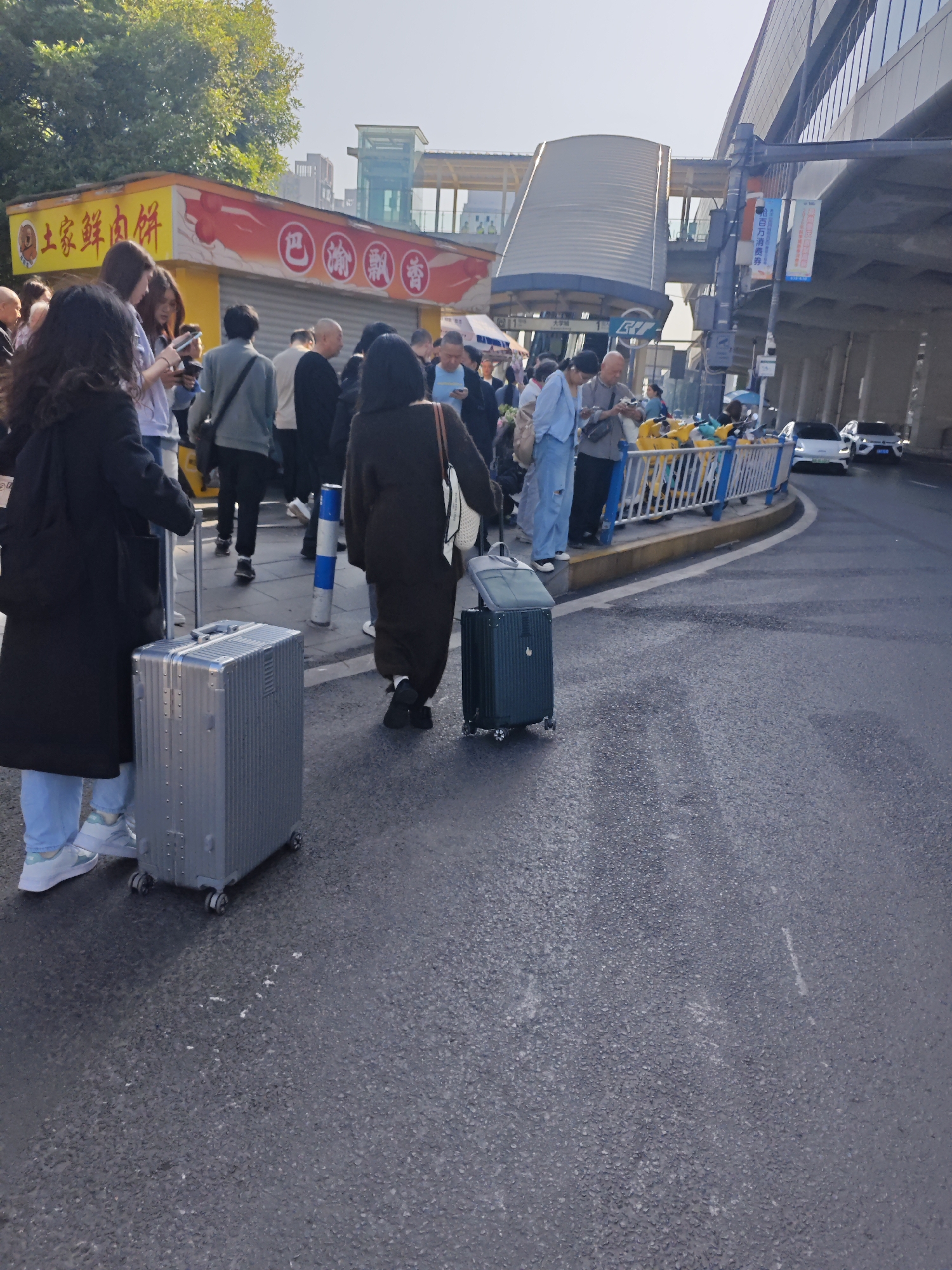The image depicts a street scene in the Chongqing High-tech Zone, China. A group of people are congregated near a street corner. Two individuals are prominent in the foreground, each with a suitcase. The person on the left has a silver suitcase and is wearing a dark coat and light blue pants. The person on the right has a dark green suitcase and is wearing a dark, full-length garment. Behind them, more people are walking and standing. A yellow food stall with Chinese writing is visible in the background. Across the street, there are parked scooters, a blue and white barrier, and two white vehicles on the road. An elevated pedestrian walkway and a building with advertisements are also in the backdrop. The ground is paved, and the lighting suggests it is daytime.