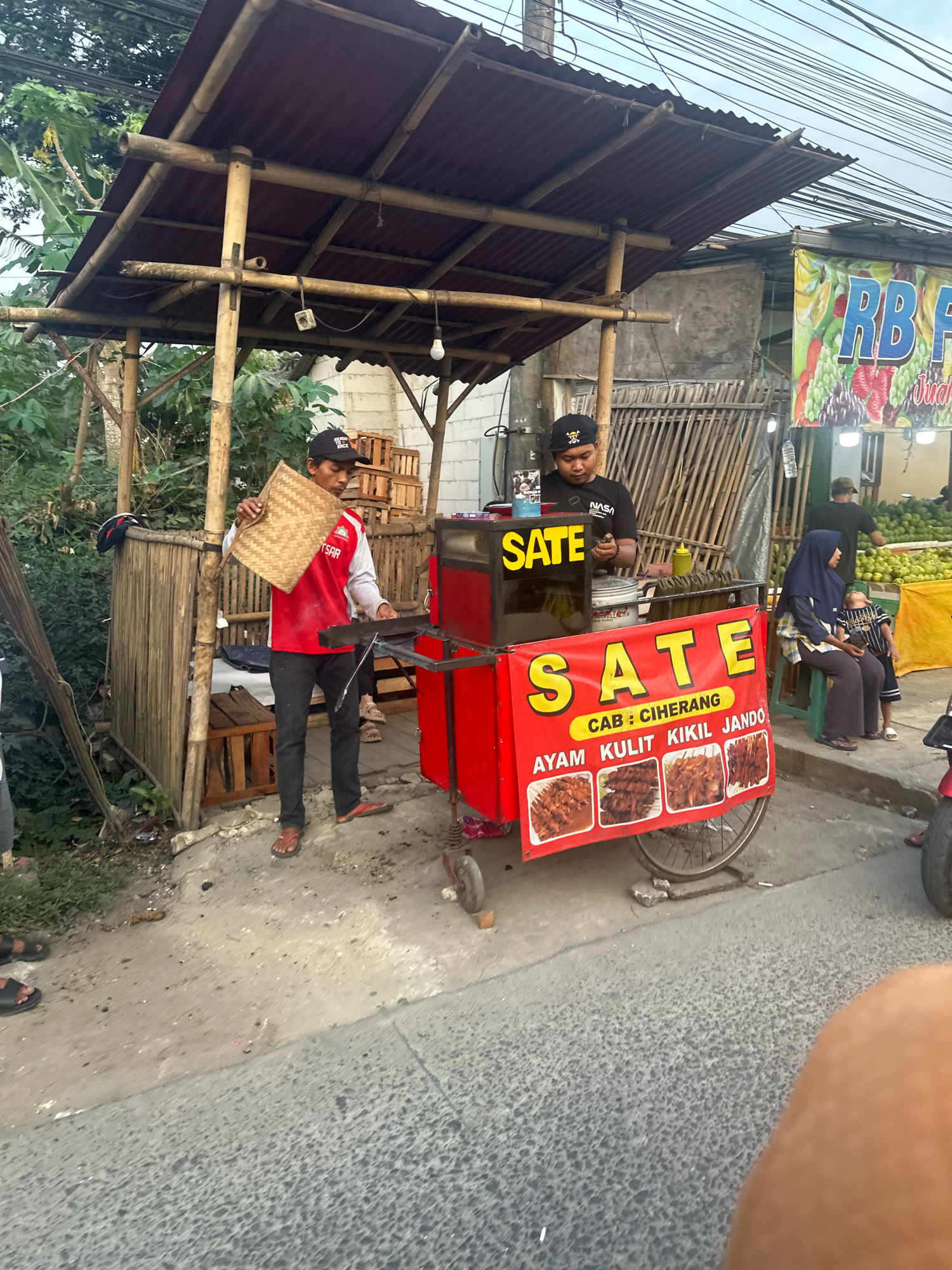 Two male vendors are operating a red food cart labeled "SATE" on a street in Saruni, Indonesia. One vendor, wearing a black baseball cap and a black t-shirt with "NASA" printed on it, is attending to the cart. He is seen looking down at the cart's contents. The other vendor, in a red long-sleeved shirt and black pants, holds a woven straw mat and appears to be preparing to serve or clean. The food cart is mobile, mounted on a bicycle wheel, and displays images of various sate dishes with Indonesian text. Behind the vendors, a bamboo-constructed stall with a corrugated metal roof provides shade. A banner with "RB FRUITS" and images of produce is visible in the background, suggesting a market or roadside vendor area. Several other people, including a woman and child seated on a bench and another person standing, are present in the vicinity. The environment appears to be an outdoor street setting with vegetation and power lines overhead. The ground is a mix of concrete and asphalt.