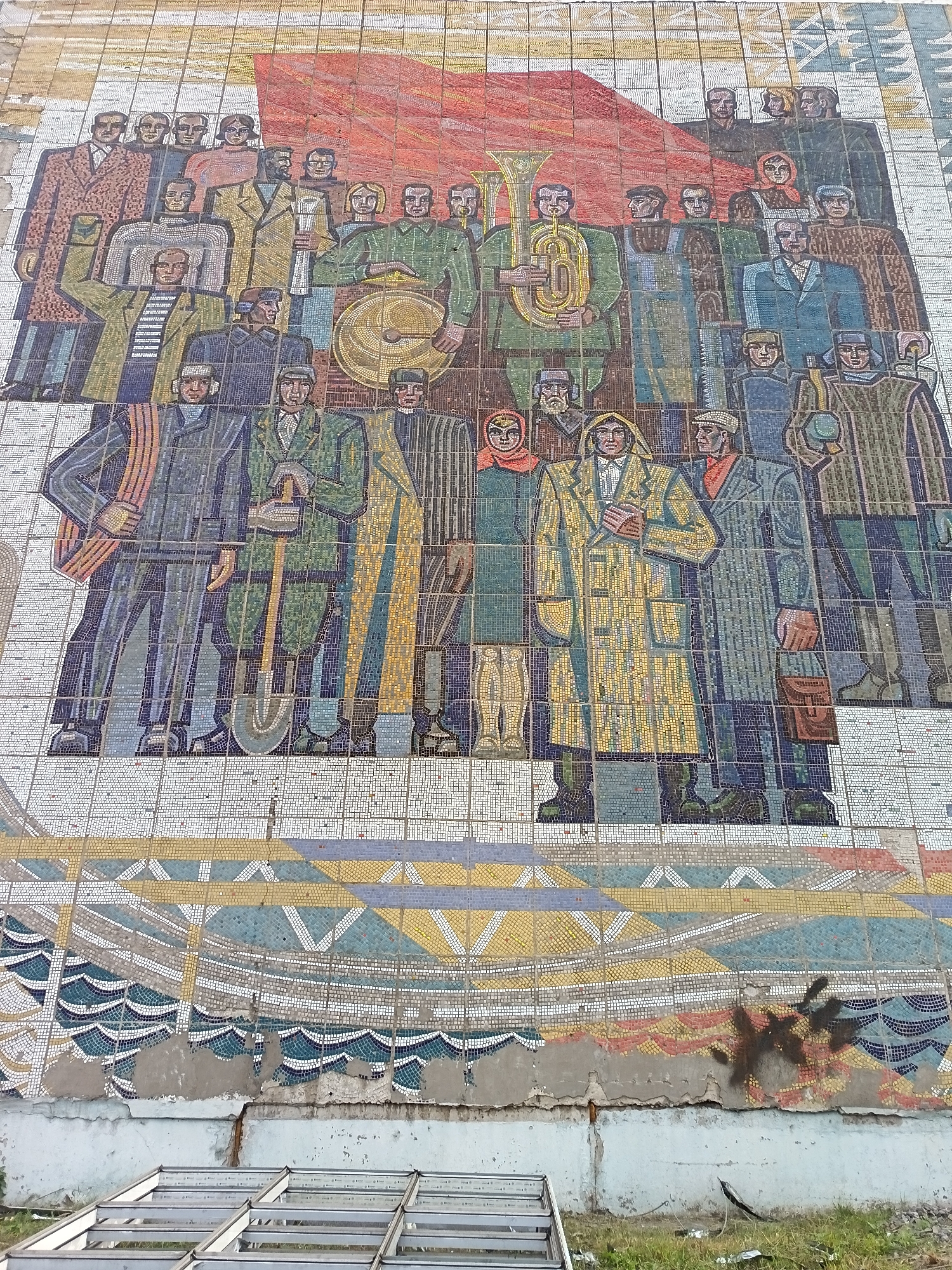 This is a mosaic mural depicting a group of people, likely in a celebratory or gathering context, set against a stylized red flag. The figures are rendered in a mosaic style with vibrant colors, primarily blues, greens, yellows, and browns, against a white and patterned background.
The subjects include men and women of various ages, dressed in what appear to be work clothes and formal attire, suggesting a diverse cross-section of society. Some individuals are holding tools such as shovels, and one person is playing a tuba, implying a connection to labor and possibly music or a parade. The overall composition conveys a sense of unity and collective spirit.
The scene appears to be outdoors, given the natural light implied by the mosaic's coloration. The lower portion of the mural transitions into abstract patterns of waves and geometric shapes, possibly representing water or a landscape element. The mural is mounted on a building, and the lower sections show signs of damage and decay, with chipped tiles and crumbling plaster. There is also graffiti present on the damaged part of the wall. Metal framing structures are visible in the foreground, suggesting ongoing construction or renovation.
There is no visible text within the mosaic itself. The style of the artwork and the imagery of people working and gathering, along with the prominent red flag, are indicative of Soviet-era propaganda art, often found in public spaces in Russia. The context of Komsomolsk-on-Amur, a city with a strong industrial history, further supports this interpretation. The emotions conveyed are generally positive, with an emphasis on strength, unity, and collective achievement.