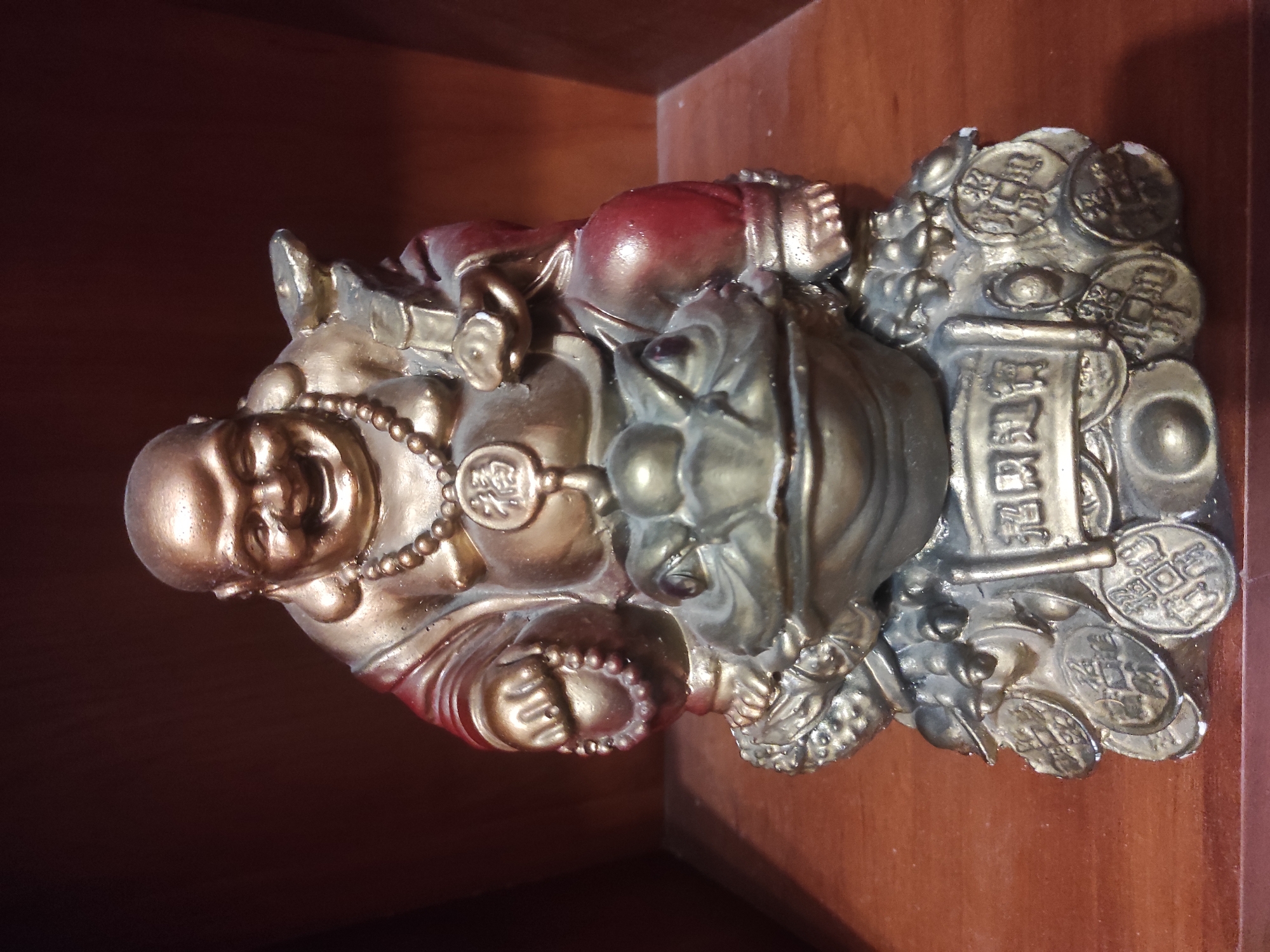 The image features a gilded statue of a laughing Buddha (Hotei or Budai) accompanied by a three-legged money toad (Chāncú), both symbolic of prosperity. The Buddha, rendered in a golden-bronze finish with parts of his robe painted red, has a wide, jovial smile and a large belly. He wears a beaded necklace with a circular medallion.
The statue is intricately detailed with various elements associated with wealth. Numerous ancient Chinese coins, some identifiable as "乾隆通寶" (Qianlong Tongbao) and "康熙通寶" (Kangxi Tongbao), are scattered around the base and near the money toad. A prominent rectangular tablet or ingot is positioned next to the toad, bearing the Chinese characters "招財進寶" (Zhāo cái jìn bǎo), which translates to "ushering in wealth and treasures." The money toad itself has a coin in its mouth, a traditional feature.
The statue is placed on a smooth, dark wooden surface, likely a shelf or table, against an equally dark wooden background, indicating an indoor setting. The lighting is gentle, emphasizing the metallic luster of the statue. There is no activity depicted; it is a static display of a decorative and auspicious item. The overall composition conveys themes of good fortune, happiness, and material abundance.