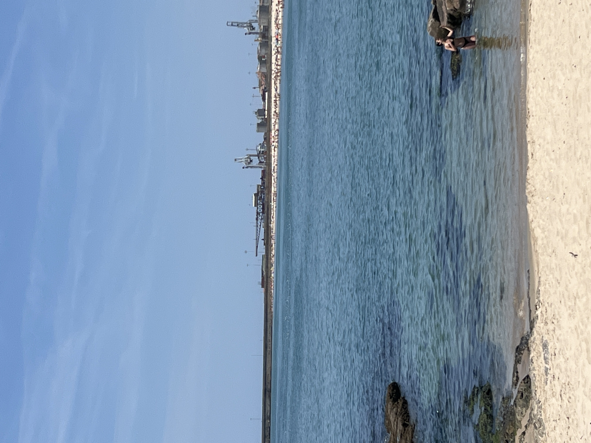 The image depicts a serene coastal scene with a woman standing in shallow, clear blue water near a sandy beach. In the background, a bustling beach with numerous sunbathers stretches along the coastline, leading towards an industrial harbor with large cranes and structures. The sky above is a clear, bright blue, hinting at a sunny day. The foreground features rocks and the sandy edge of the beach. The overall impression is one of a vibrant, accessible coastline where leisure and industry coexist, likely in the vicinity of Porto, Portugal's port area.