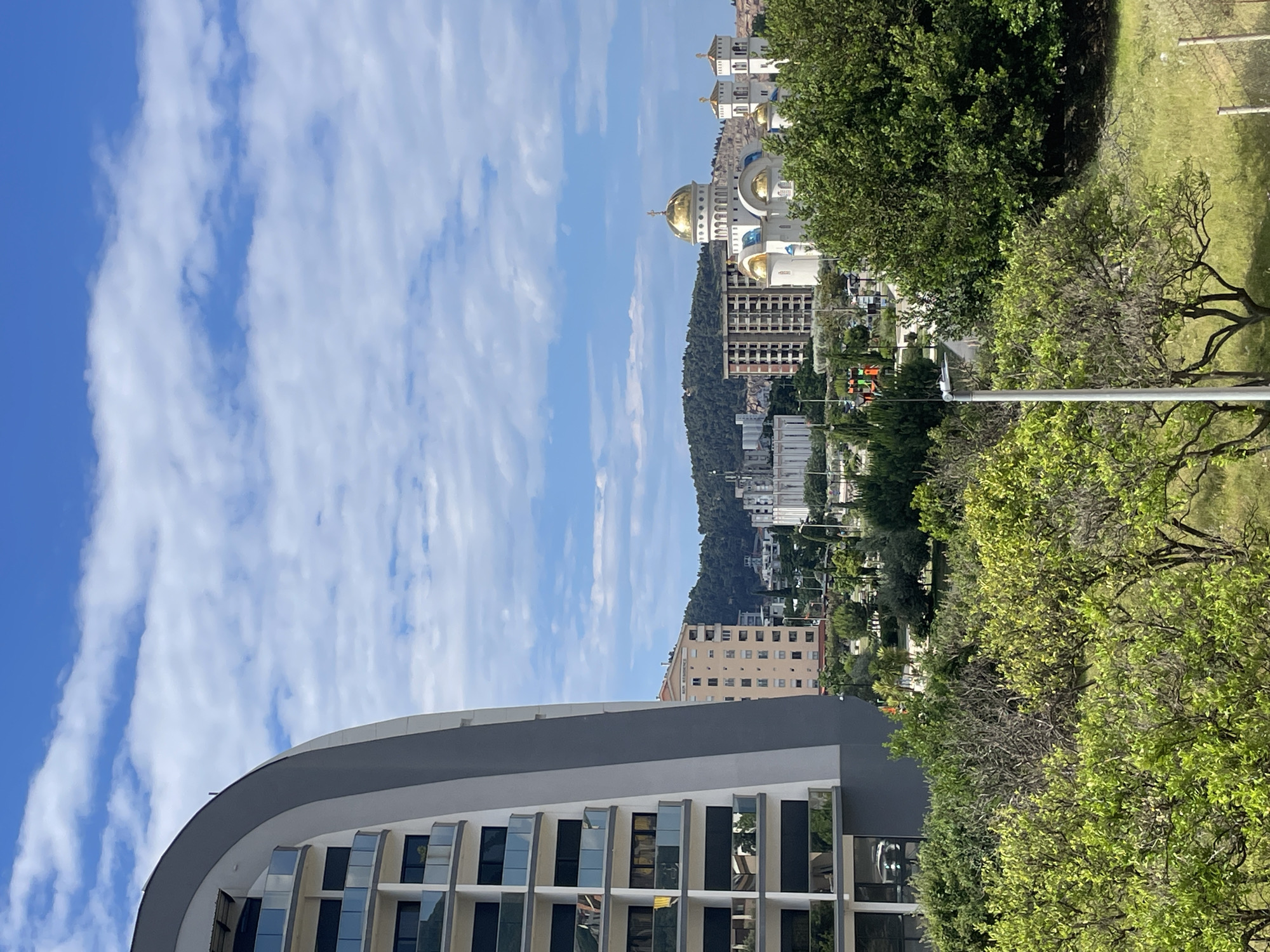 The image depicts a cityscape in Bar, Montenegro, under a blue sky with scattered white clouds. In the foreground, a modern building with a curved roof and reflective windows is partially visible. Behind it, a densely populated area with various buildings, including residential structures and a prominent Orthodox church with golden domes, is present. The church appears to be atop a slight hill. Trees and greenery are interspersed throughout the scene, adding natural elements to the urban landscape. A utility pole is visible on the right side of the frame.