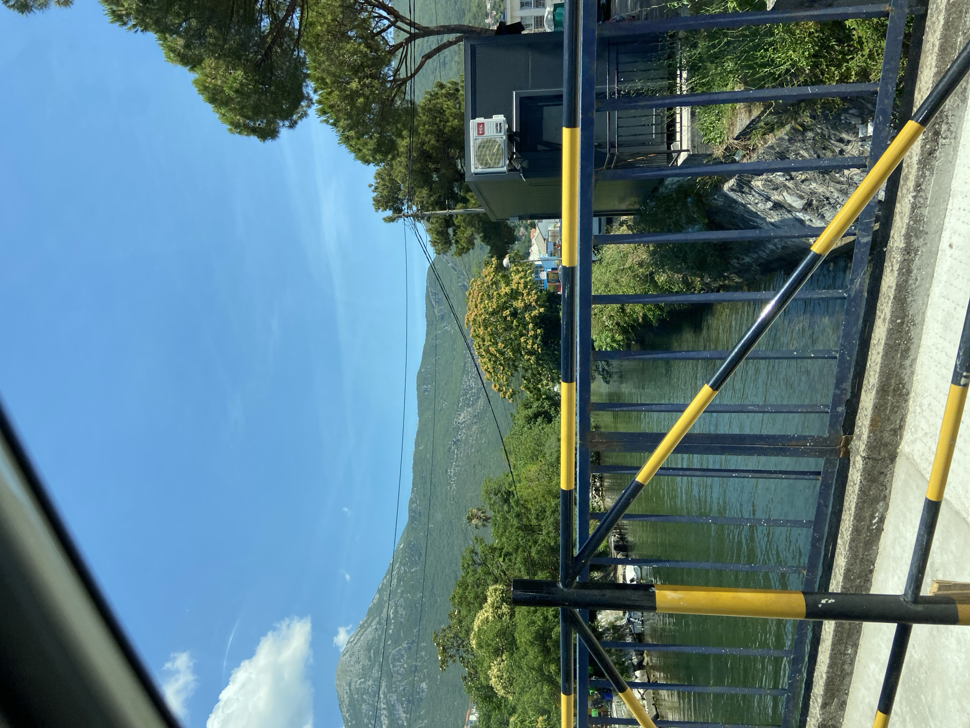 This is a daytime, outdoor, eye-level shot taken from a bridge overlooking a canal or river. The bridge railing, painted black with yellow stripes, is in the foreground. The water in the canal is a deep green and appears calm with reflections. Beyond the canal, lush green trees and foliage line the banks. In the mid-ground, a dark gray building with a TCL air conditioning unit mounted on it is visible. Further back, a large, green, tree-covered mountain dominates the landscape, with a clear blue sky above dotted with a few wispy white clouds. The scene is set in Zelenika, Montenegro.