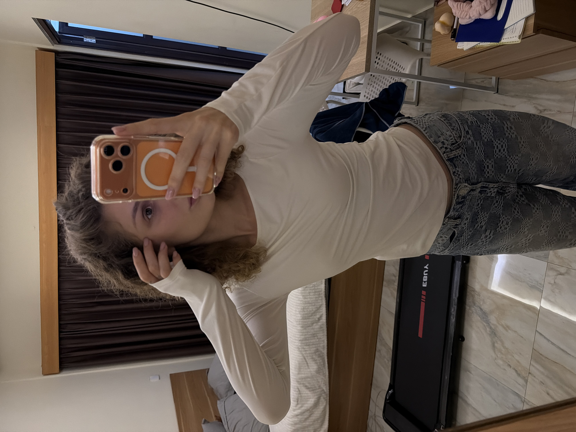 A female with curly hair is taking a selfie in a room. She is wearing a white long-sleeved shirt and patterned jeans. She is holding an orange phone case in her hand, partially obscuring her face. The room contains a bed, a desk with items like a headband, and a folded treadmill. The room is located in North Kuta, Indonesia.