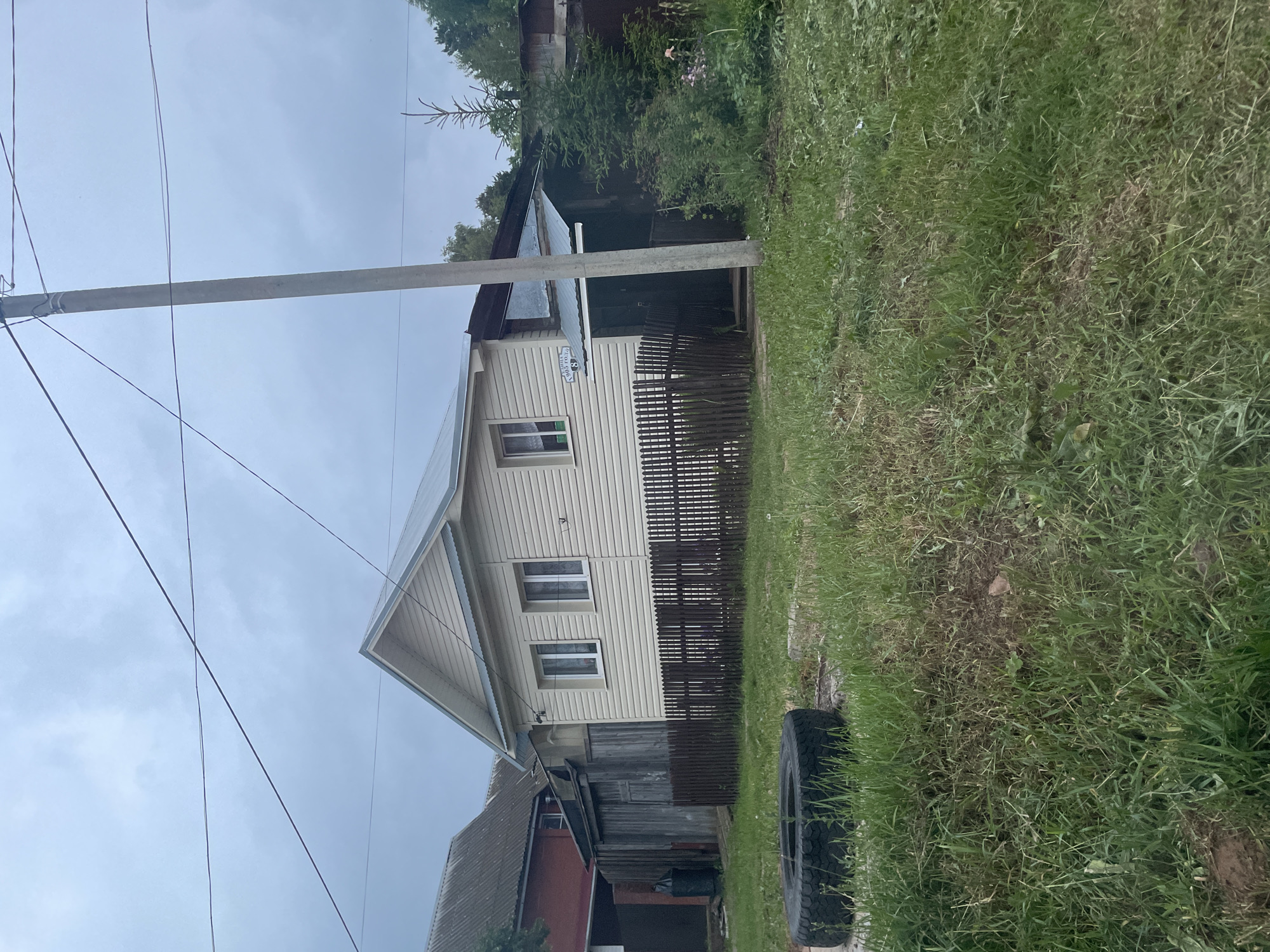 The image captures a static, outdoor scene of a residential property on an overcast day, located at 82 Ulitsa 905 Goda (Street of the Year 1905) in Votkinsk, Udmurt Republic, Russia.
The central subject is a single-story house featuring light cream-colored horizontal siding and a grey metal gable roof. Two white-framed rectangular windows with white curtains are visible on the facade. To the left of the rightmost window, a blue oval sign is affixed, displaying the street name "УЛИЦА 905 ГОДА" and the house number "82" in white text. A dark brown wooden picket fence, approximately hip-height, extends across the front of the property. To the right of the main house, a covered entrance or porch with a corrugated metal roof is partially obscured by a grey concrete utility pole.
To the left and slightly behind the primary house, another building with a dark, possibly red or brown, exterior and a grey corrugated roof is visible. A wooden fence or outbuilding structure connects the two houses.
In the immediate foreground, the ground is covered with unkempt green grass, showing patches of bare soil. A large, black vehicle tire, possibly from a truck or agricultural vehicle, lies flat on the grass in the left portion of the foreground.
A tall, grey concrete utility pole stands prominently in the right-center of the frame, extending from the ground to the top edge. Multiple black utility lines traverse the sky, connected to the pole. The sky is uniformly light grey, indicating overcast conditions. Trees and dense green foliage are visible in the background, particularly to the right of the property. No individuals or active subjects are present in the scene.