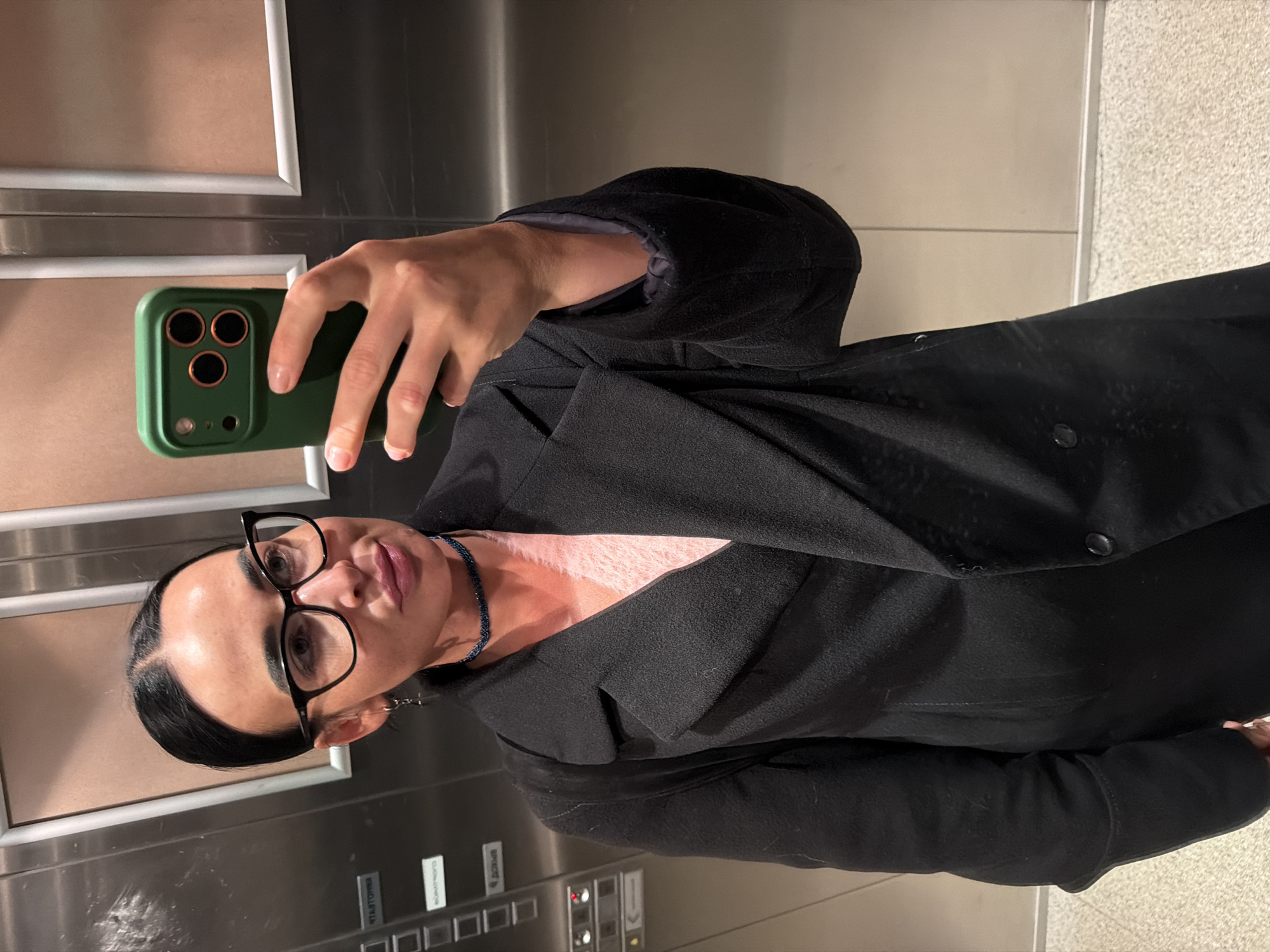 A person with dark hair and glasses is taking a selfie with a green smartphone in what appears to be an elevator. The person is wearing a black coat. The elevator buttons have inscriptions in Russian. The location is likely in Saint Petersburg, Russia, given the Cyrillic script on the buttons.
