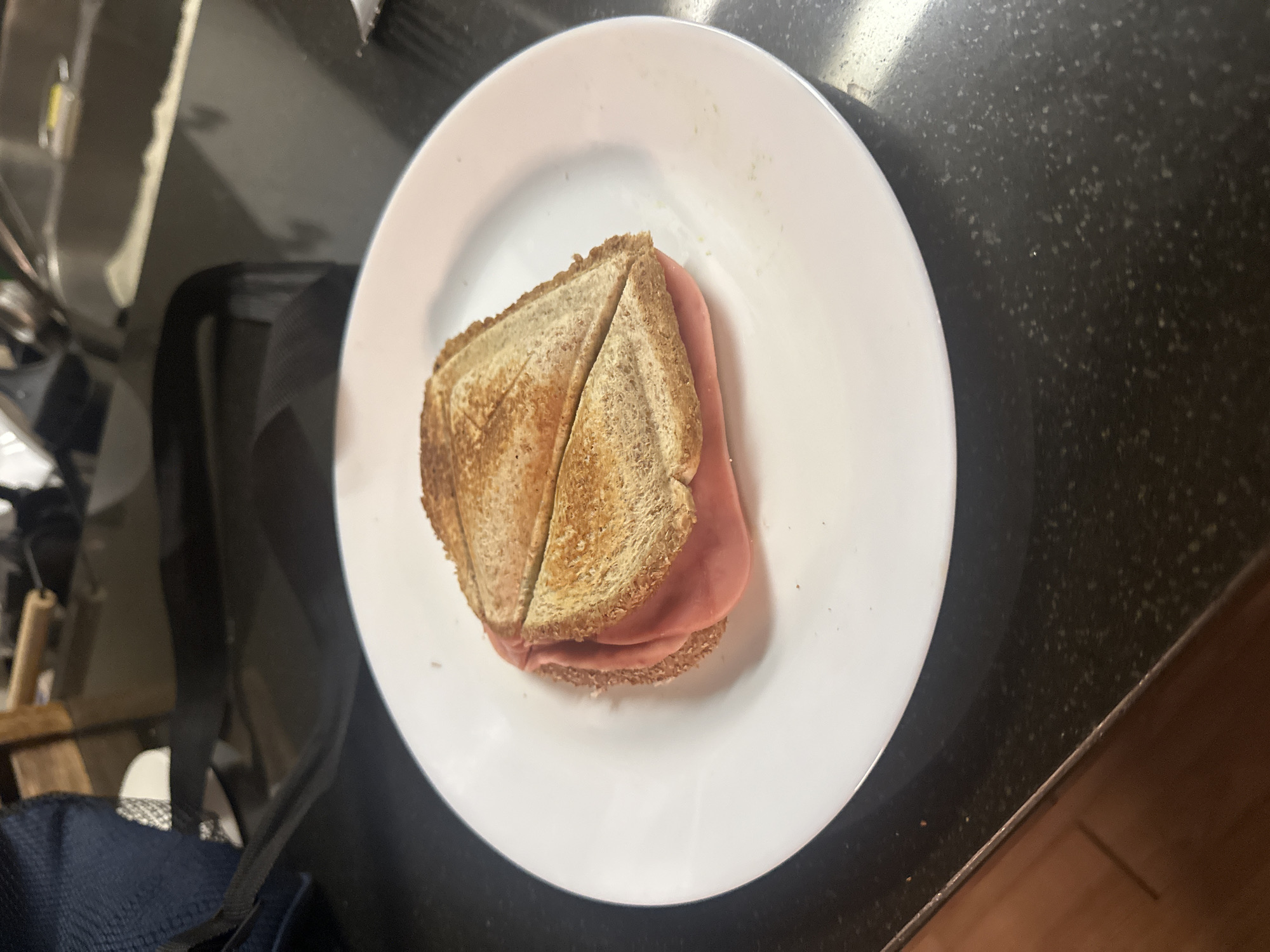 A sandwich is placed on a white plate. The sandwich is cut in half diagonally and consists of toasted bread and ham. The plate is on a dark countertop. In the background, a sink and various kitchen items are visible. The overall setting appears to be a kitchen environment, possibly in a residential area of Huixquilucan de Degollado, Mexico, given the request. There are no people or animals visible in the image.