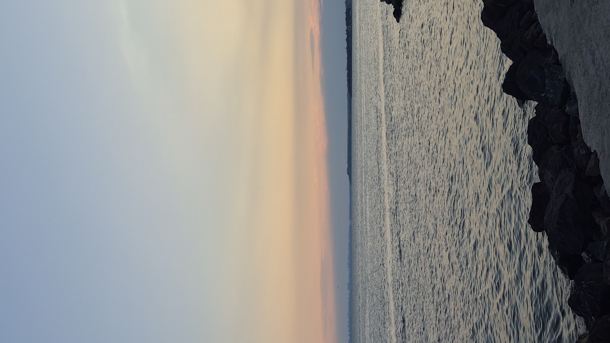 A vertical, full shot captures a serene sunset over the Black Sea at Pomorie, Bulgaria. The sky transitions from a pale blue at the top to soft hues of orange and pink near the horizon, where a distant city skyline is faintly visible. The calm sea reflects the muted colors of the sky, with gentle ripples disturbing its surface. In the foreground, dark, jagged rocks line the edge of the water, with a concrete pathway visible on the right. No people or animals are present in the scene. The overall impression is one of peaceful tranquility at the end of the day.