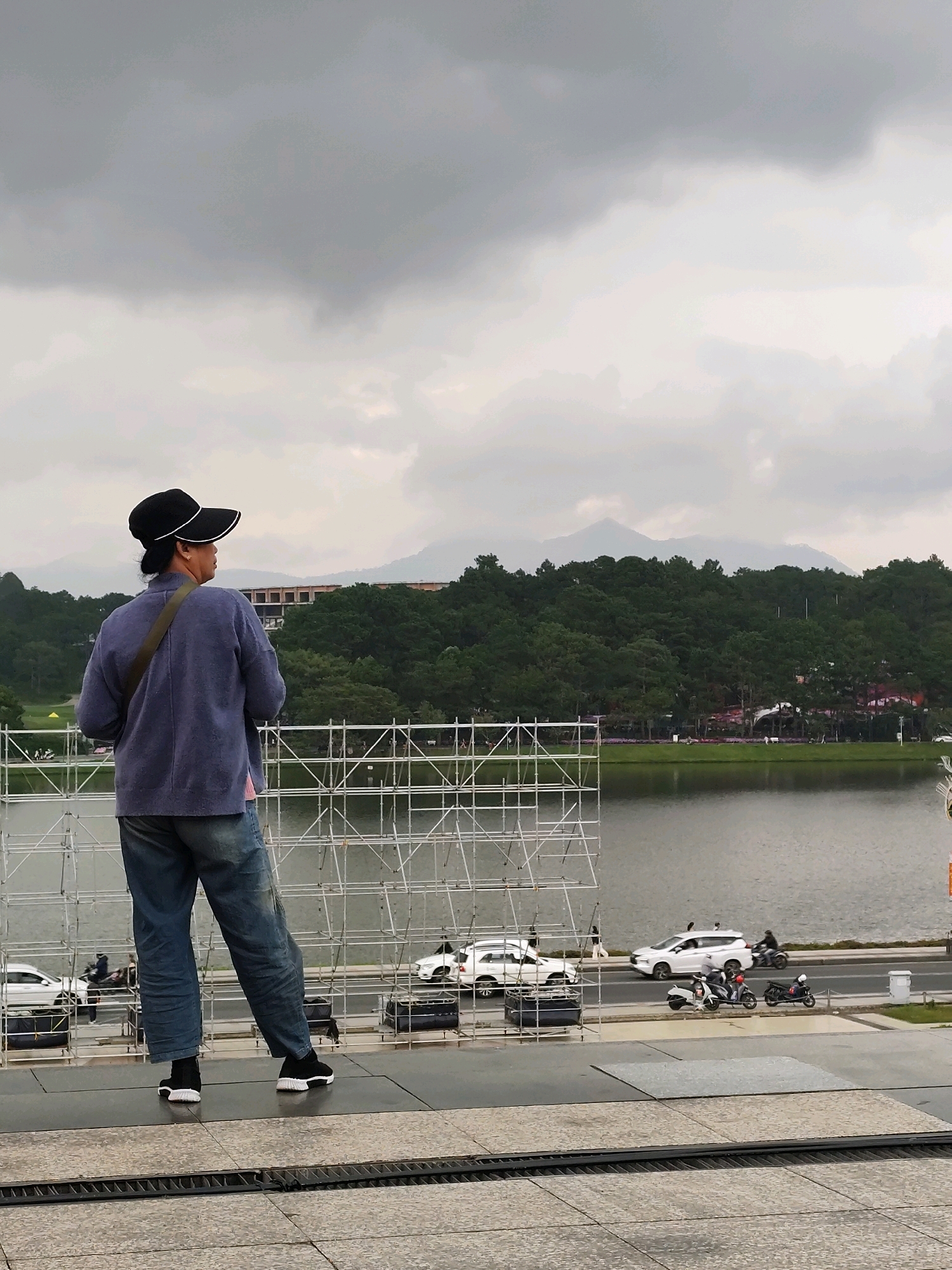 A person wearing a blue sweater, jeans, and a black hat stands facing away from the camera, looking out over a body of water. In the background, there is a lush green forest with mountains in the distance under a cloudy sky. Along the water's edge, there are roads with cars and motorcycles. In the foreground, there is scaffolding and a paved walkway. This scene is likely the Xuan Huong Lake area in Da Lat, Vietnam, a popular tourist destination known for its picturesque landscape and recreational activities.
