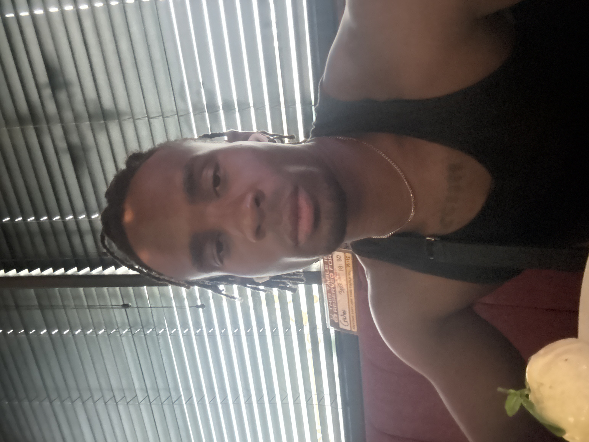A selfie shows a Black male, wearing a black tank top and gold necklace, with braided hair. Visible on his left collarbone is a tattoo. He is sitting in a booth at a restaurant in Unknown, Unknown. The window behind him has Venetian blinds. A sign on the table reads "This table is reserved". Part of a salad is visible on the table.