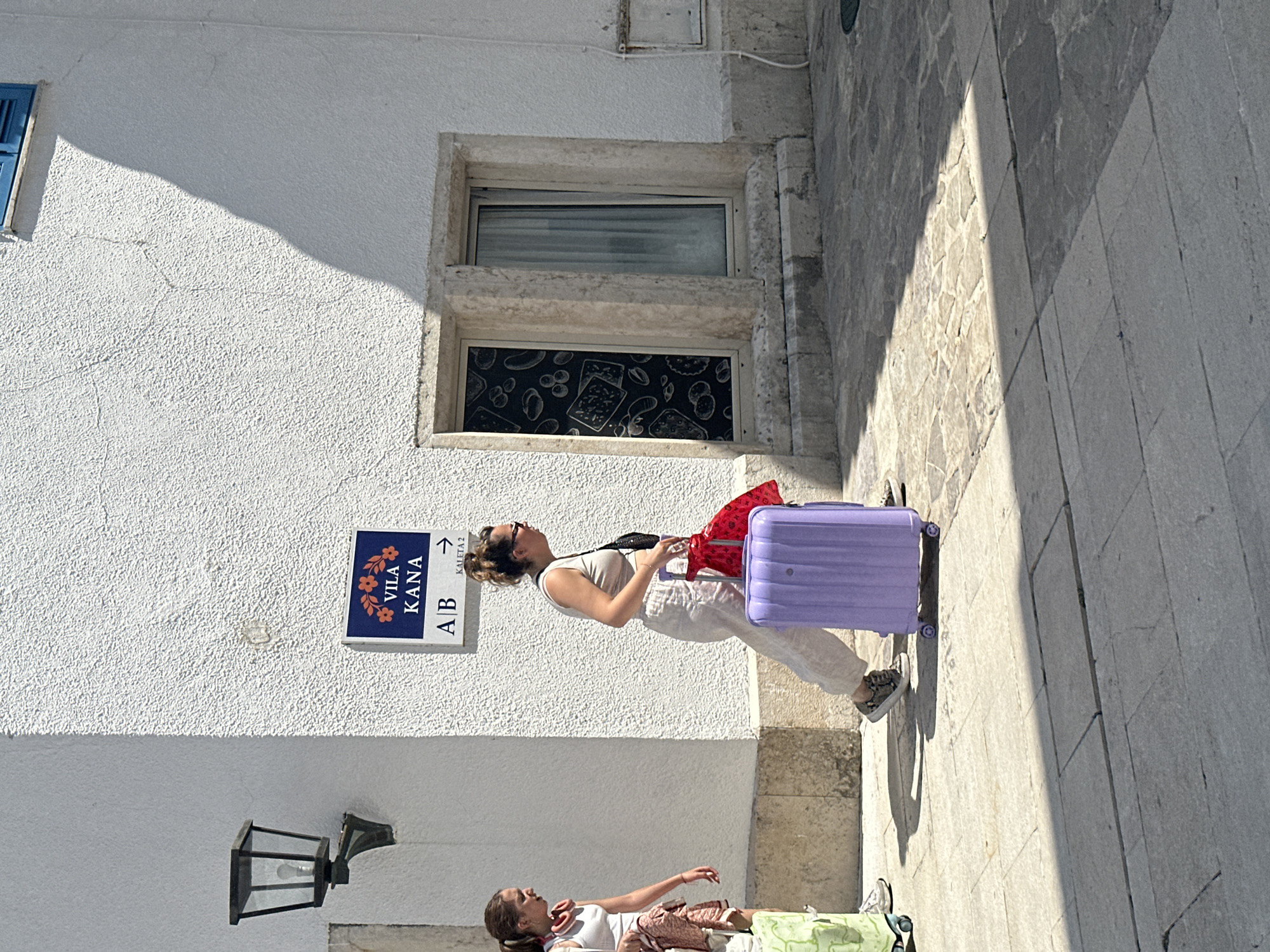 In Budva, Montenegro, on a bright, sunny day, two women are captured in motion. The woman in the foreground, on the right, is walking forward and slightly to the right, pulling a lavender suitcase with four wheels. She is wearing a light-colored tank top, loose-fitting trousers, and patterned sneakers. A black crossbody bag is slung over her shoulder, and she holds a red cloth bag in her left hand. Her hair is pulled back in a bun. To her left, the second woman is walking towards the left side of the frame. She has her hair in a ponytail and wears a white tank top and shorts. She is carrying a light-colored bag over her left shoulder and appears to have another bag draped over her arm. The scene is set on a cobblestone street in front of a textured, white building. A sign with a floral motif and the text "VILA KANA" is visible on the building's exterior, along with directional arrows. A black lantern is mounted on the wall to the left of the sign. Two windows are visible in the building; one has a decorative pattern visible inside, and the other has a sheer curtain. The sunlight casts strong shadows on the ground, indicating a high sun position.
