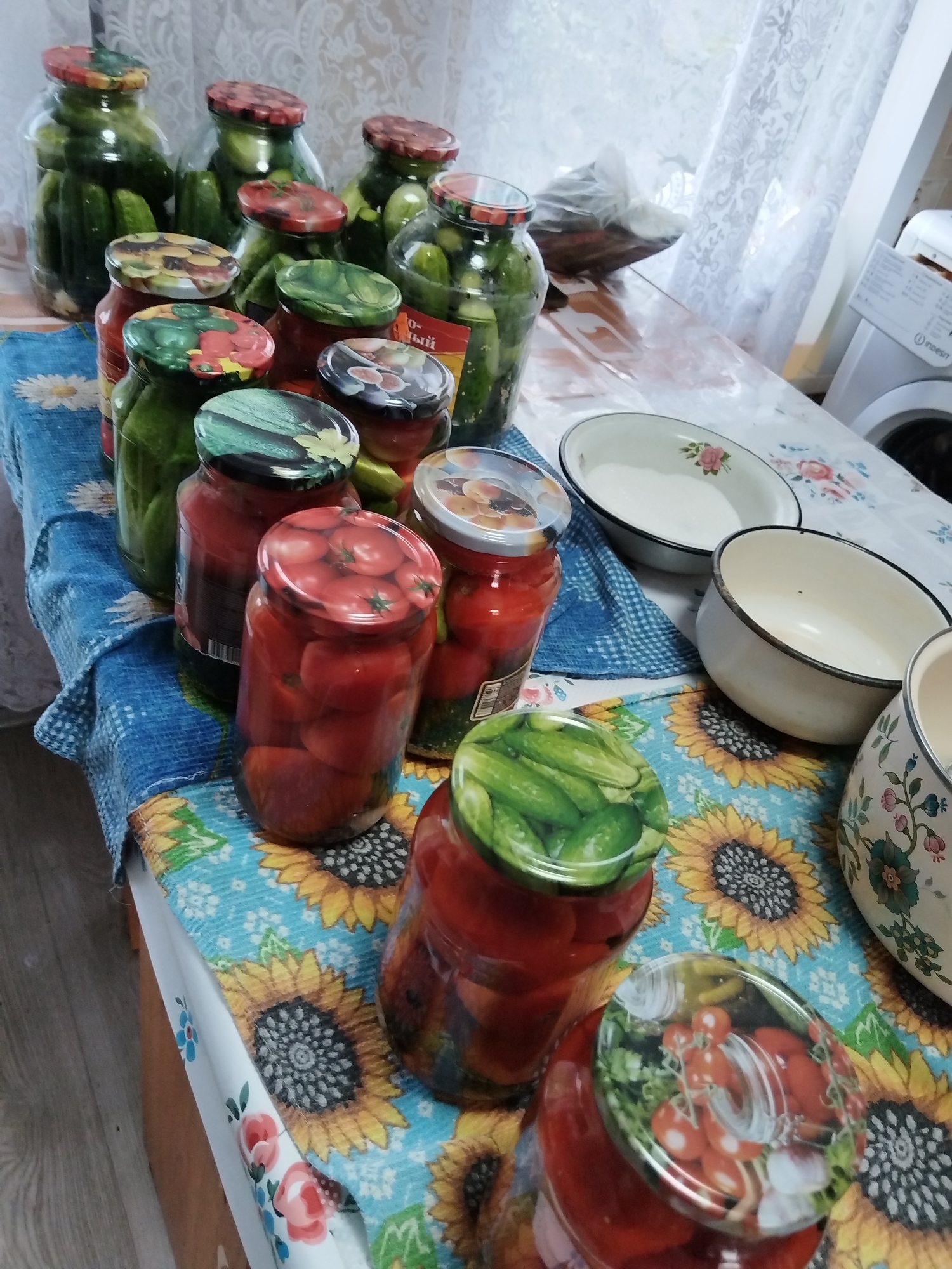 The image shows a table laden with jars of preserved food. There are several jars of pickles and tomatoes visible. The table is covered with a blue tablecloth featuring a sunflower pattern. In the background, a white washing machine is partially visible. The setting appears to be a domestic kitchen or pantry in Minayevka, Russia. There are no people in the image, and no actions are taking place. The scene is static and depicts preserved food items ready for storage.