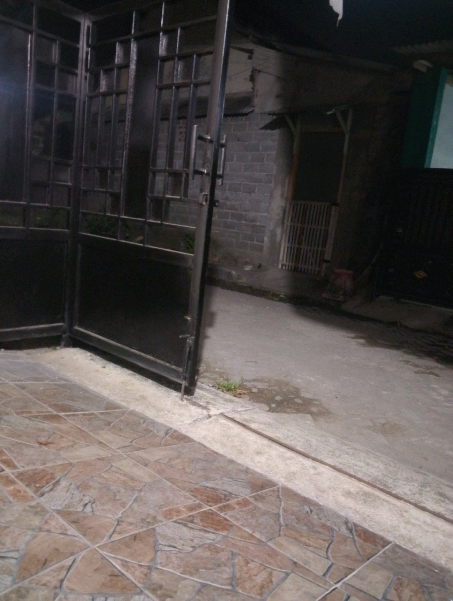 The image shows a dark, outdoor scene. In the foreground, there is a tiled floor with a mosaic pattern of brown and beige stones. To the left and center, there is a large, black metal gate with a grid pattern. Part of the gate is open, revealing more of its structure. Behind the gate, the environment appears to be an alleyway or a backyard. The walls are constructed of grey bricks. Further back, there is a doorway with a slatted wooden gate and a dark interior. To the right of the image, there is a concrete ground with some puddles of water, suggesting it may have rained recently. In the distance, other structures and fences are visible, all shrouded in shadow due to the low light conditions. The scene gives the impression of being in a residential area or a back alley in the city of Kendalrejo, Indonesia, likely at night or during very low light conditions. There are no people or animals visible in the image.