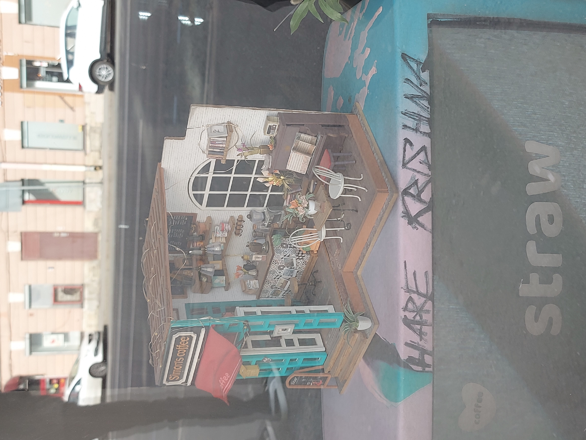 The image captures a miniature diorama of "Simon's coffee," a coffee shop, displayed in what appears to be a storefront window. The diorama features a detailed interior with miniature furniture, coffee-making equipment, and a sign indicating "Simon's coffee." Outside the cafe, there is a red awning with the word "coffee" on it. The diorama is set against the backdrop of a city street visible through the window. The name "Hare Krishna" is written in black marker on the container holding the diorama. A larger sign "straw coffee" is also visible. The reflection in the window shows the exterior of buildings and vehicles. The location appears to be Saint Petersburg, Russia, based on the architectural style and the presence of the "Simon's coffee" sign, which is a known cafe in the city.