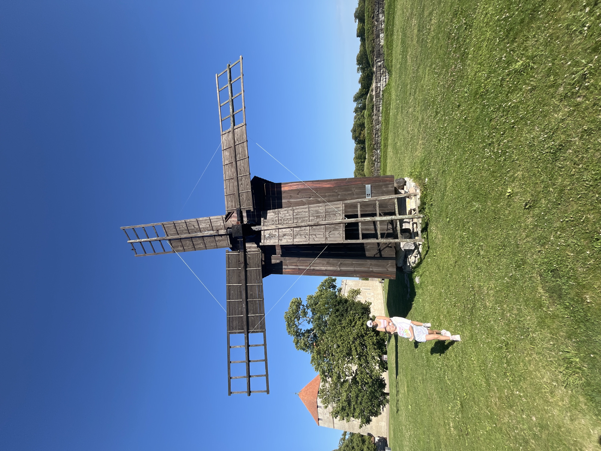 The image shows a large wooden windmill with four blades extended against a clear blue sky. In front of the windmill, two people, a woman and a child, are standing on a green grassy field. The woman is wearing a white top and pink shorts, and the child is wearing a white t-shirt and patterned shorts. Behind them, a stone tower with a red roof and a large green tree are visible. The scene appears to be set in a historical park or museum. The location is Kuressaare, Estonia, specifically in the courtyard of Kuressaare Castle.