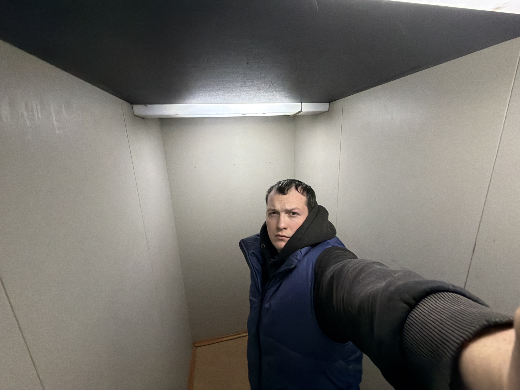 A man stands in what appears to be a small, enclosed space, possibly an elevator or a very small room. The walls are light grey, and the ceiling is dark. There is a fluorescent light fixture on the ceiling. The man is wearing a dark hooded sweatshirt and a blue puffer vest with dark sleeves. The image seems to be a selfie taken at an upward angle. The bottom of a wooden structure is visible at the base of the wall on the left. There is no indication of location within Chaikovsky, Russia.