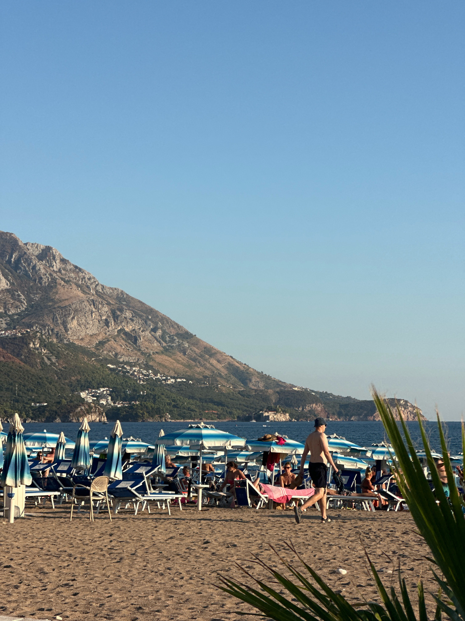 A man wearing a blue cap and black shorts walks across a sandy beach in front of rows of blue and white striped beach umbrellas and lounge chairs. The man is partially silhouetted against the bright sky. In the background, the blue water of the Adriatic Sea meets a mountainous coastline dotted with white buildings. Several people are seated on lounge chairs under the umbrellas, some with towels. The overall scene depicts a sunny day at a popular beach resort.