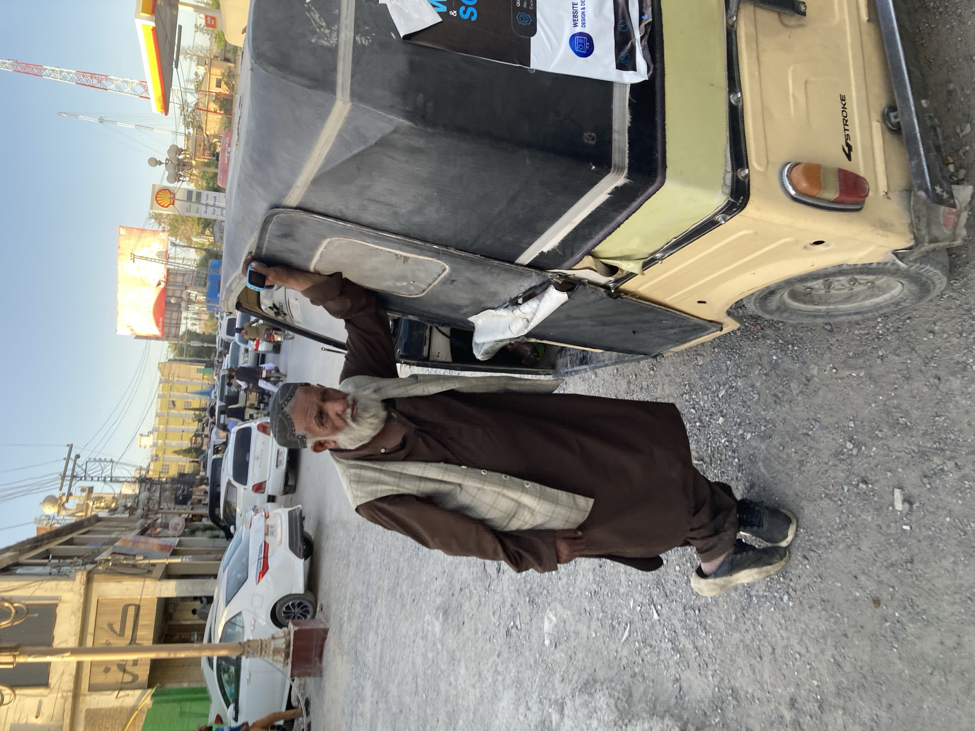 A man stands next to an auto-rickshaw on a street in Quetta City Tehsil, Pakistan. He wears a brown robe with a patterned vest, and black shoes. The auto-rickshaw is light yellow with a black roof. In the background, there are buildings, cars, and utility poles. The sky is clear and the sun is shining.
