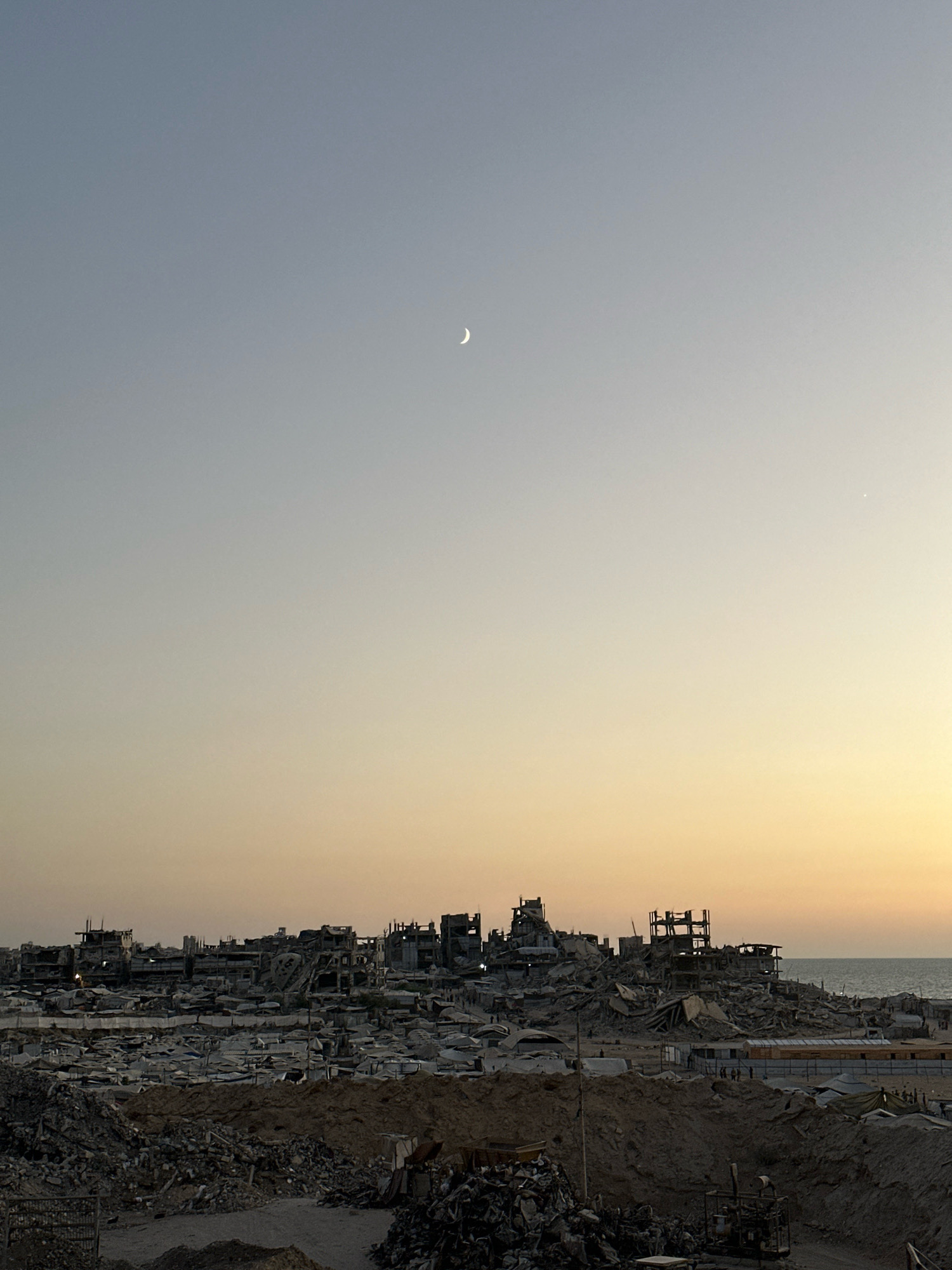 The image depicts a panoramic view of widespread destruction in a densely populated urban area, identified as the city of Gaza, Palestinian Territories. The scene is characterized by the ruins of numerous buildings, reduced to rubble and skeletal structures of rebar and concrete. Debris piles are prominent in the foreground and spread throughout the landscape, indicating extensive damage.
In the midground, rows of damaged structures and what appear to be makeshift shelters or tents are visible, densely packed together. The remnants of larger buildings, some with exposed internal frameworks, dominate the skyline. The overall impression is of a war-torn environment.
In the distance, the calm sea is visible, contrasting with the devastation on land. The sky displays a gradient of colors, transitioning from a clear pale blue at the top to warmer hues of orange and yellow near the horizon, suggesting either dawn or dusk. A thin crescent moon is visible in the upper left quadrant of the sky, and a small, bright star or planet is faintly visible in the upper right quadrant.
Several individuals are faintly discernible as small figures amidst the ruins and structures, engaged in various activities, though their specific actions cannot be determined due to the distance and scale. The environment is desolate, with minimal visible vegetation. The presence of sand and dirt further contributes to the impression of a heavily impacted and potentially neglected area.