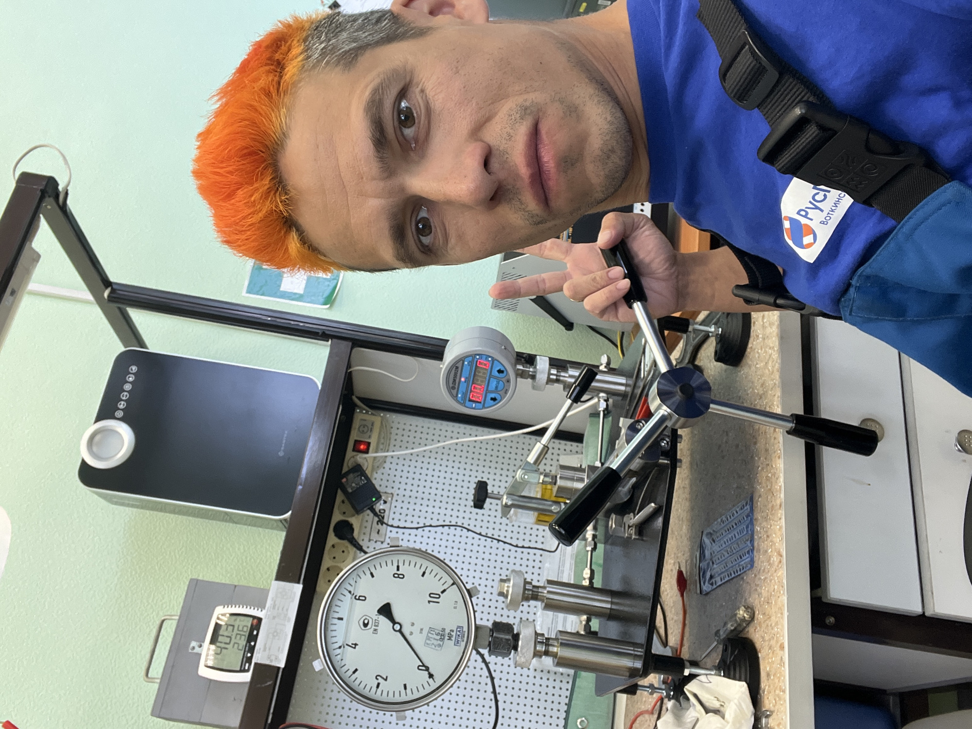 The image shows a man with dyed orange hair taking a selfie. He is wearing a blue shirt with the logo of "Pych Botkins" on it, indicating the location of Botkinsky Machine Building Plant in Chaikovsky, Russia. He is standing next to a piece of equipment with gauges and handles. One gauge is circular with a dial numbered from 0 to 10, while another is digital and shows the number 40.3. The man is holding a handle and making a peace sign with his fingers. There is also a black rectangular device with circular buttons on the left side of the frame.