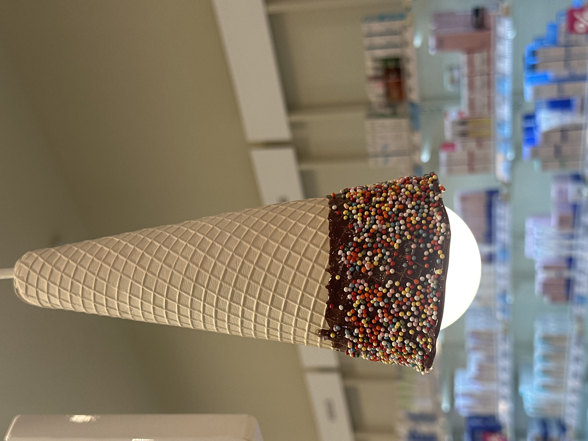 A low-angle shot depicts an ice cream cone-shaped lamp, covered in chocolate and colorful sprinkles, against a blurred background of a shop interior. The waffle cone texture is visible. The room appears to be a shop in Stavropol, Russia, characterized by shelves stocked with an assortment of products. The lamp's light is visible within the cone.