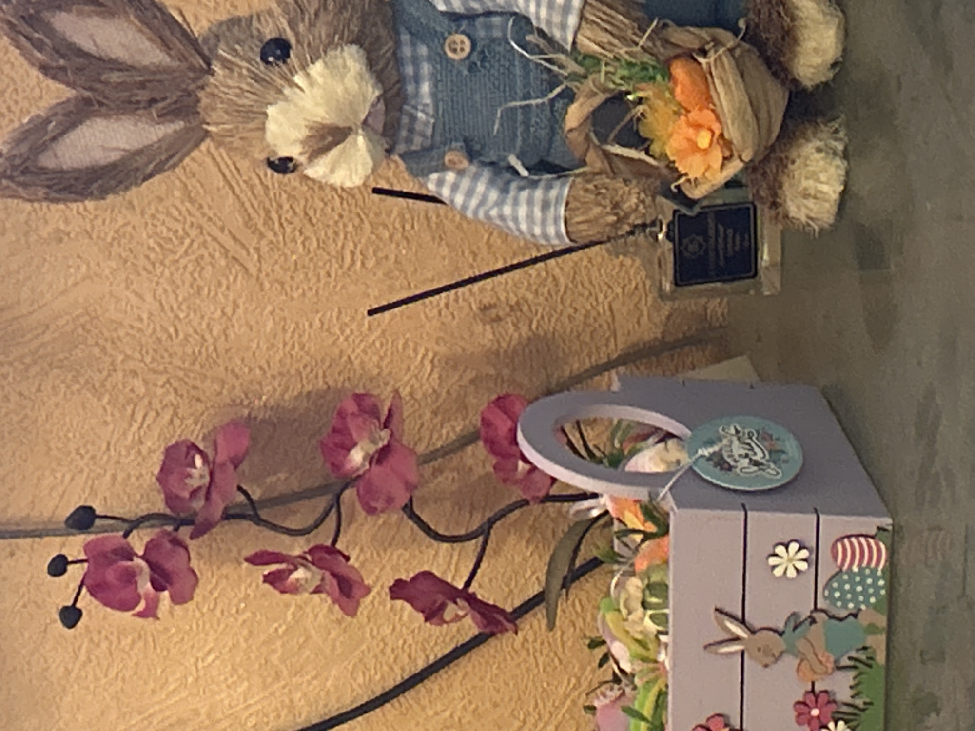 Here's a description of the image:
The image shows a decorated interior setting. There's a straw bunny doll wearing a blue and white checkered shirt with a vest on the top left. The bunny is holding a bouquet of flowers and greenery tied with burlap. Attached to the flowers is a small, dark-colored tag. Below and to the right of the bunny is a light purple wooden box, decorated with a bunny, flowers and polka dots. The box also contains an assortment of artificial flowers and foliage. A round tag with a bunny on it is hanging on the box. A decorative metal branch with dark pink orchid-like flowers is positioned diagonally between the bunny and the box. The background appears to be a textured, golden-brown wall. There is no information about the location being in Sofia, Bulgaria.