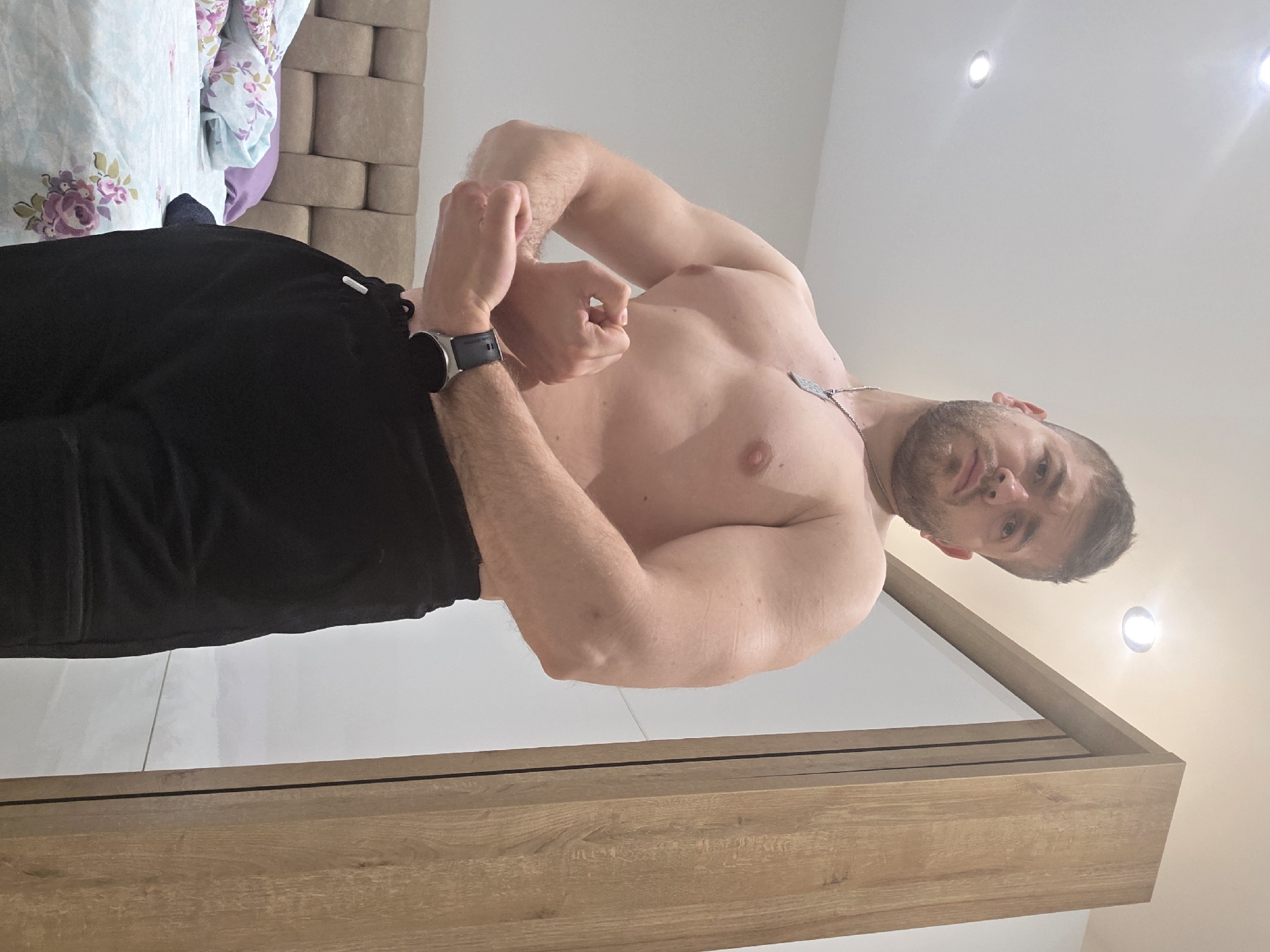 A shirtless man is standing indoors in front of a mirror, flexing his arm muscles. He is wearing black sweatpants and a black smartwatch on his left wrist. The background includes a bed with floral bedding, a headboard with a brick-like pattern, and a ceiling with recessed lighting. The location is not specified as Yagodovo, Bulgaria, in the image.