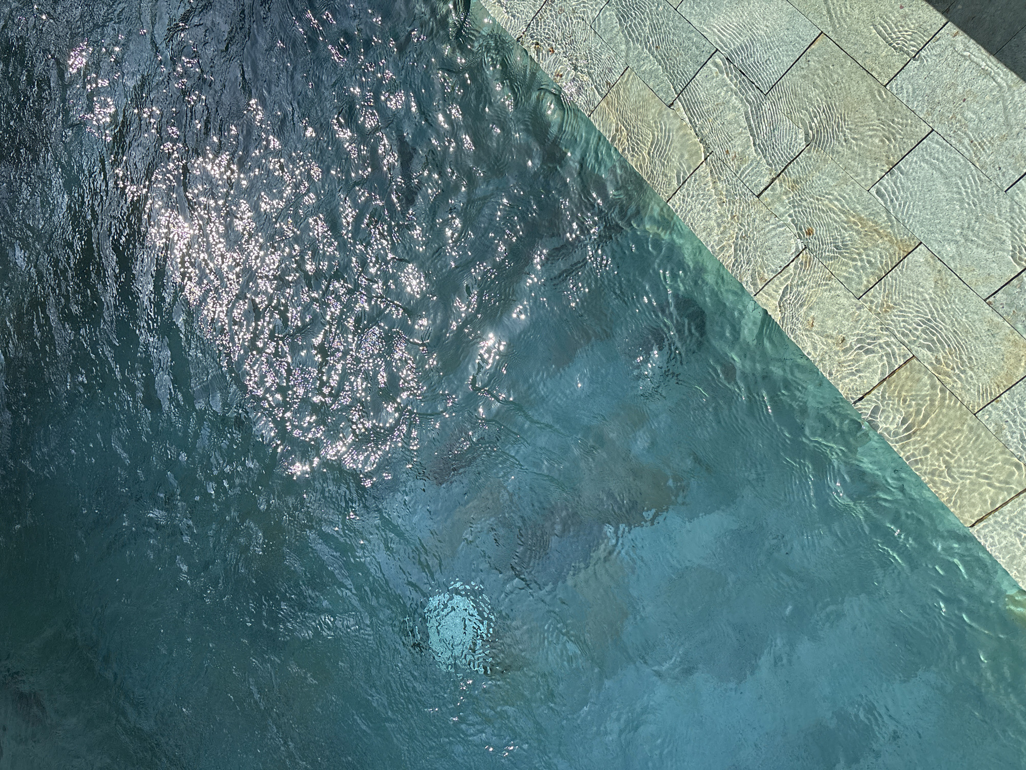 The image is a top-down view of a swimming pool, likely located in Cemagi, Indonesia. The pool water is blue-green and reflects sunlight. The pool edge is lined with light-colored stone tiles. The water's surface is rippled.