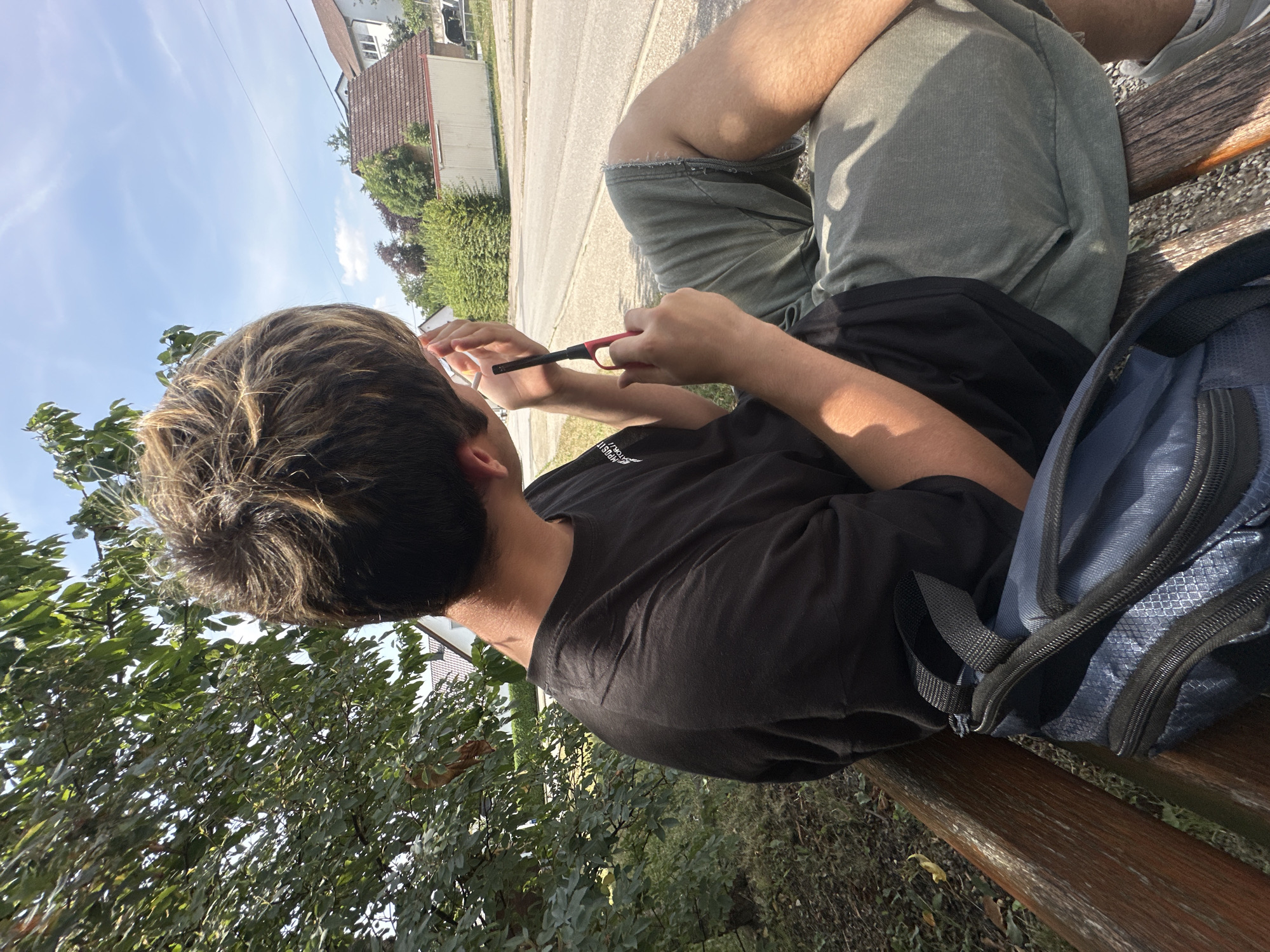 A person sits on a wooden bench in a park-like setting, looking to the right and holding a lighter and a cigarette. The person is wearing a black t-shirt and grey shorts. A blue backpack rests on the bench next to them. In the background, there is a street with houses and trees, suggesting an urban or suburban environment. The sky is blue with some clouds, and the sun is shining, casting shadows. This scene is likely located in Günzburg, Germany, given the architectural style of the houses and the overall appearance of the landscape.