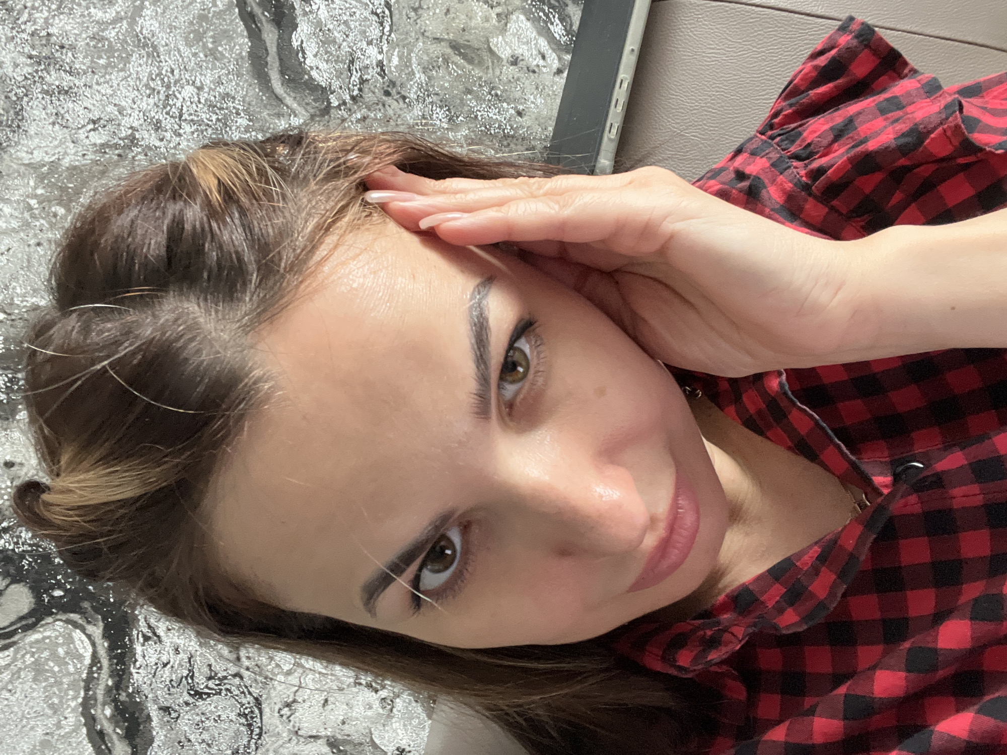 A woman with brown hair and eyes is lying down, looking up at the camera. She has her hand on her forehead. She is wearing a red and black checkered shirt. The background includes a textured, possibly marble-like surface and a beige surface that may be a couch or chair. The location is Unknown, Unknown.